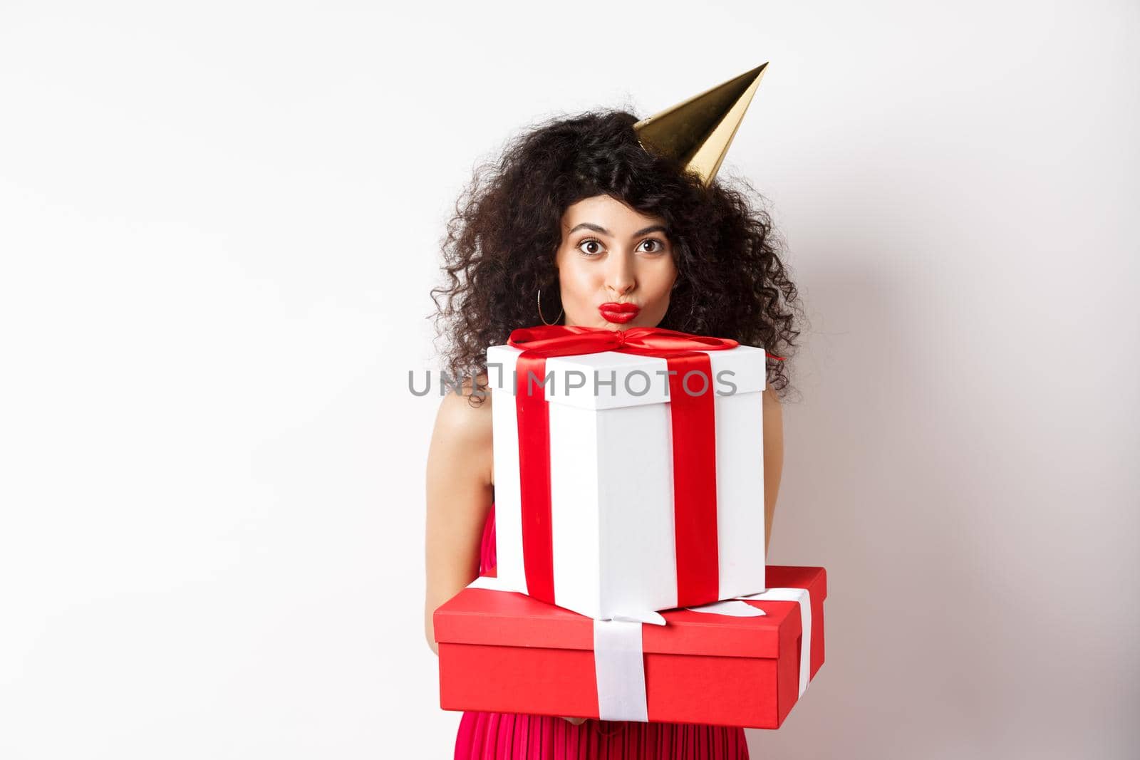 Cute birthday girl with curly hair and party hat, holding gifts and looking happy at camera, standing against white background.