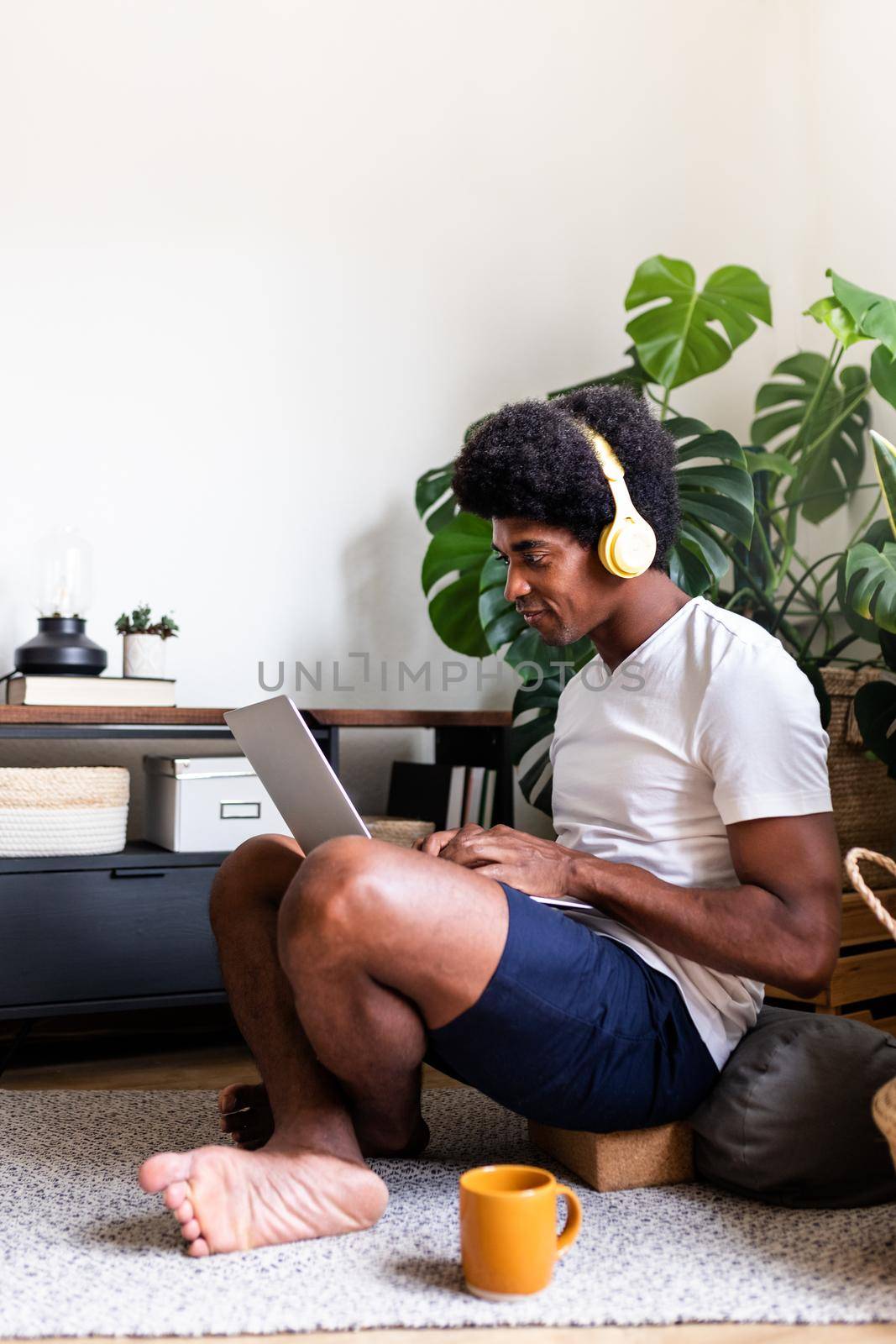 Young african american man using laptop wearing headphones in living room. Vertical image. Copy space. by Hoverstock