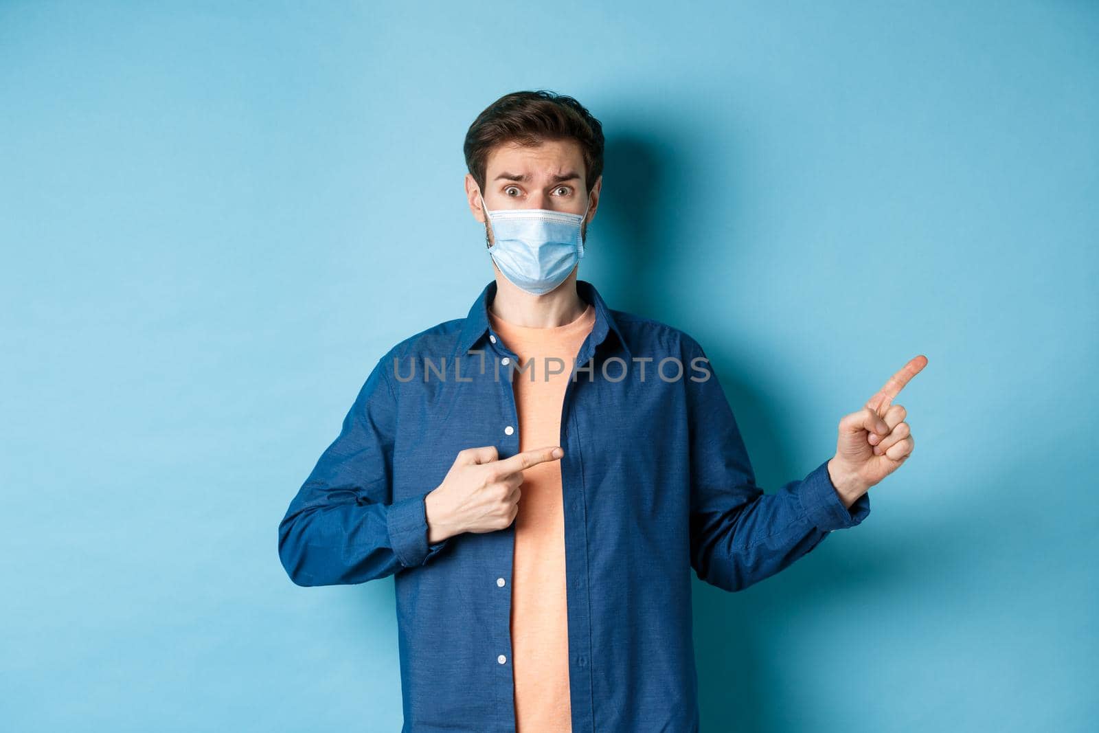 Covid and healthcare concept. Worried young man in medical mask raising eyebrow, pointing finger right with doubtful and nervous face, standing on blue background by Benzoix