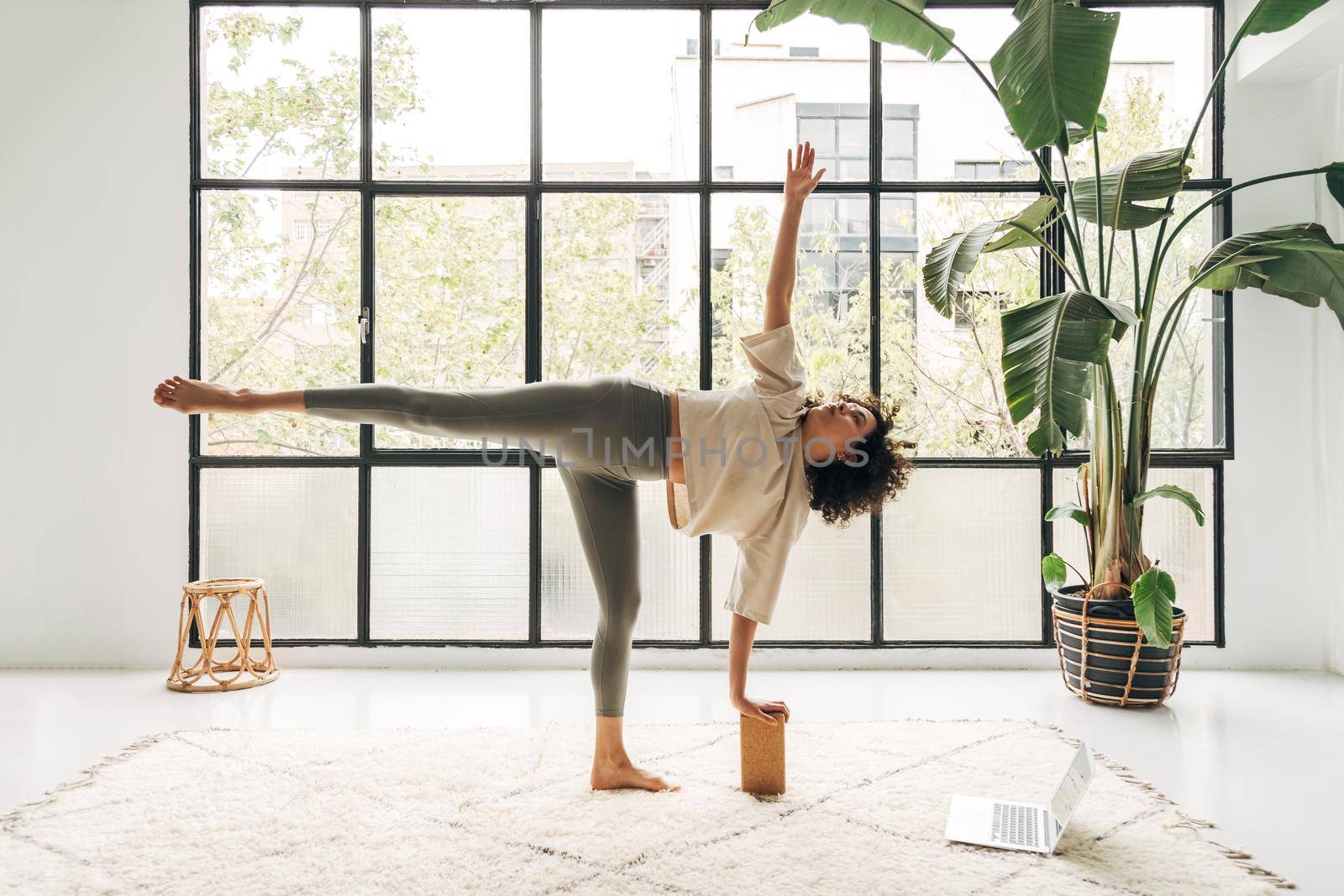 Young african american woman doing online yoga classes at home using laptop. Ardha chandrasana, half moon pose. Spirituality and active lifestyle concepts.