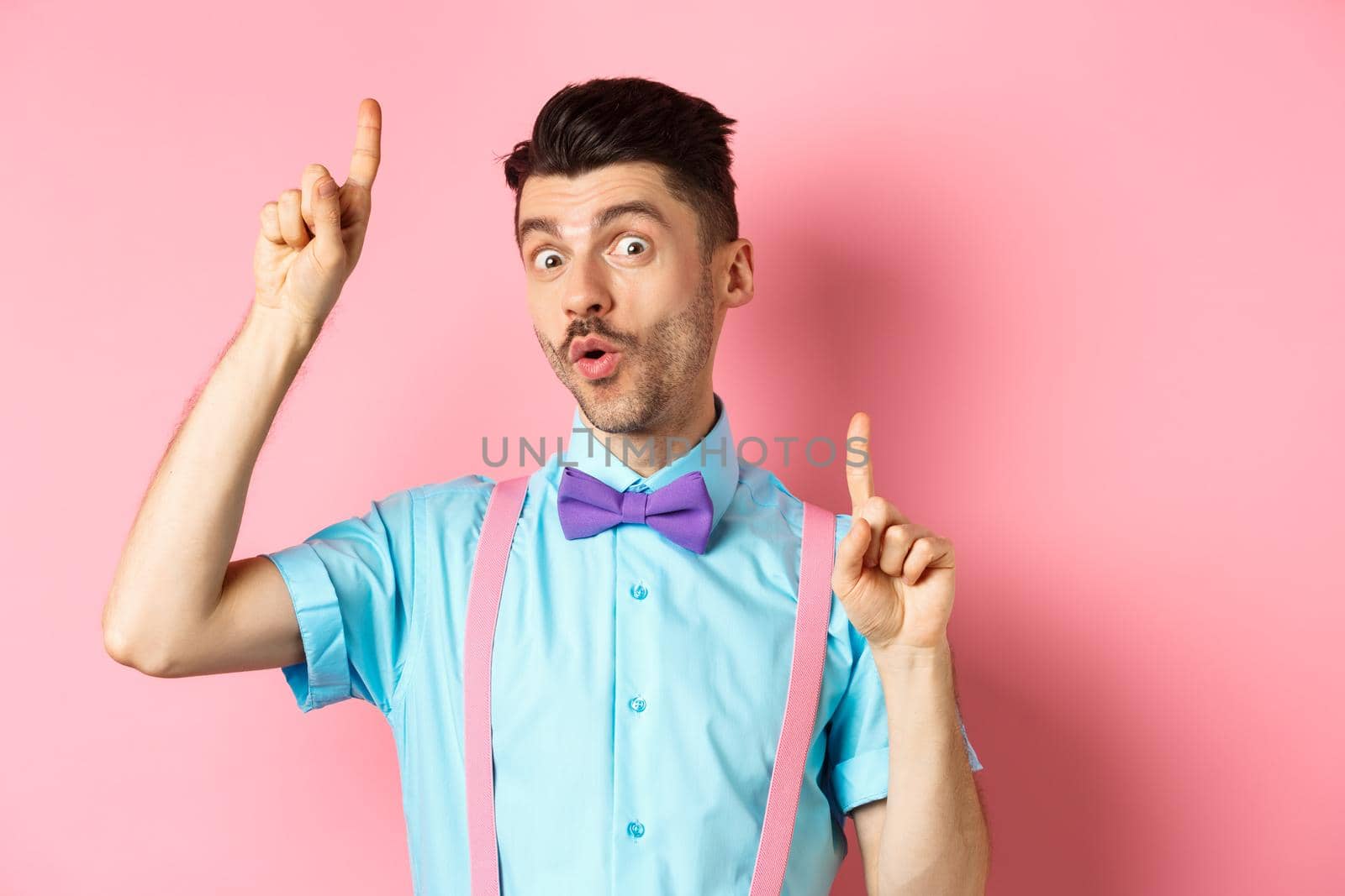 Cheerful funny man dancing in bow-tie and suspenders, pointing fingers up and looking upbeat, standing over pink background.