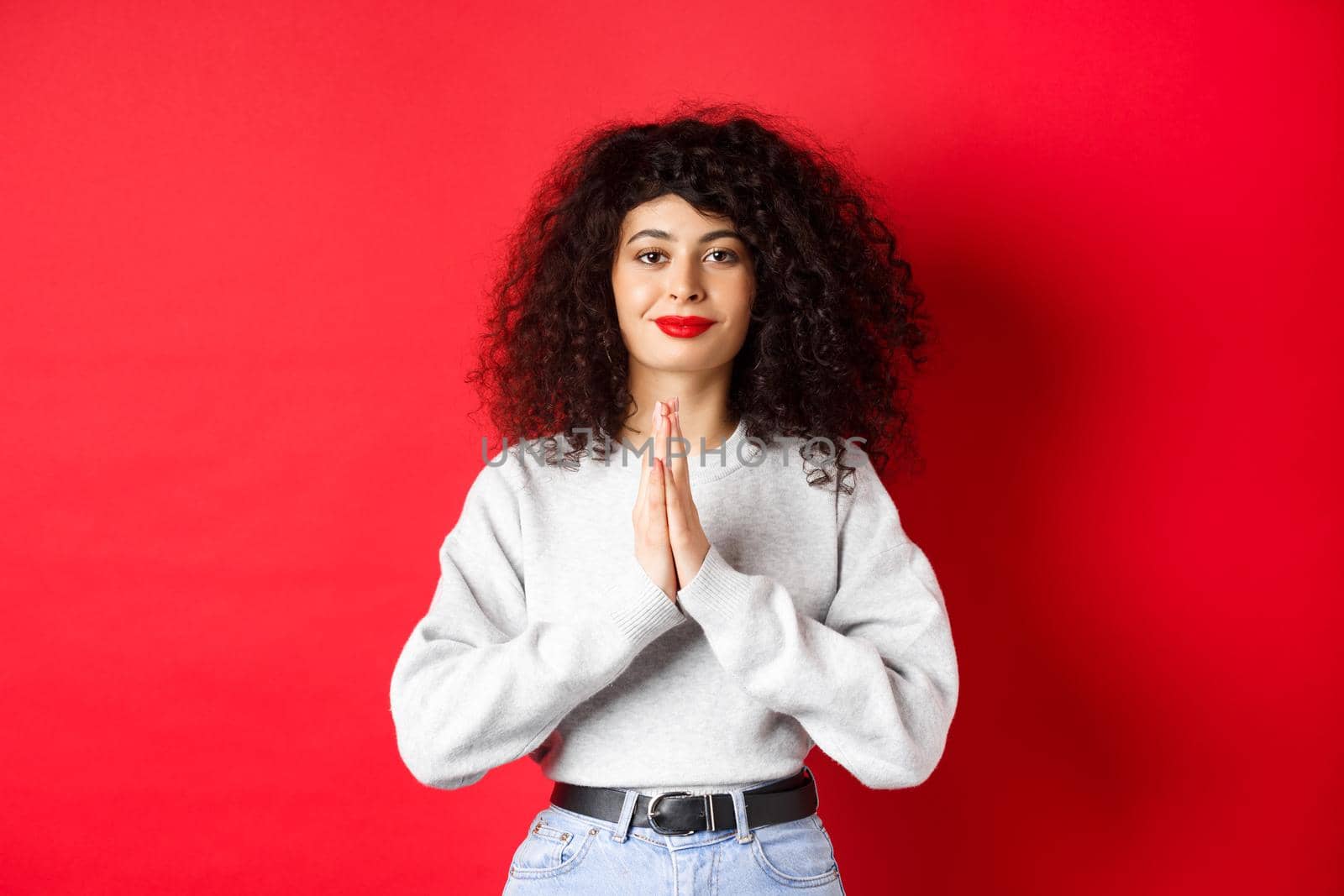 Namaste. Thankful young woman with curly hair and red lips show gratitude, smiling pleased at camera, standing on studio background by Benzoix