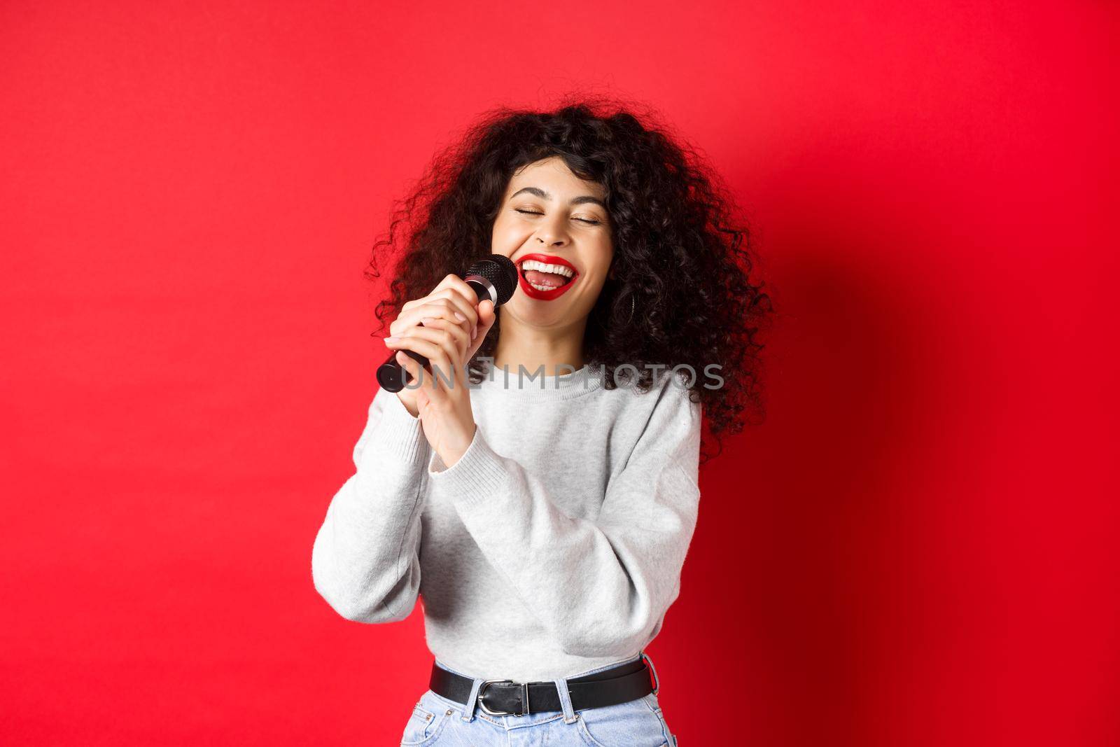 Hobbies and leisure concept. Happy woman singing song in microphone, having fun at karaoke with mic, standing on red background by Benzoix