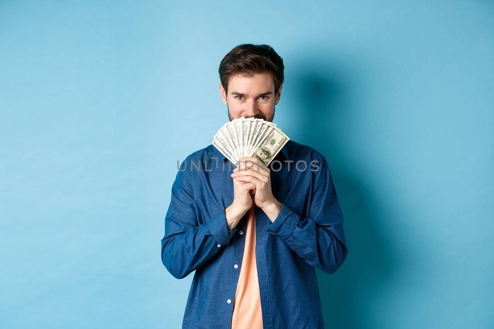 Young man hiding smiling face behind money and looking determined at camera, standing on blue background by Benzoix