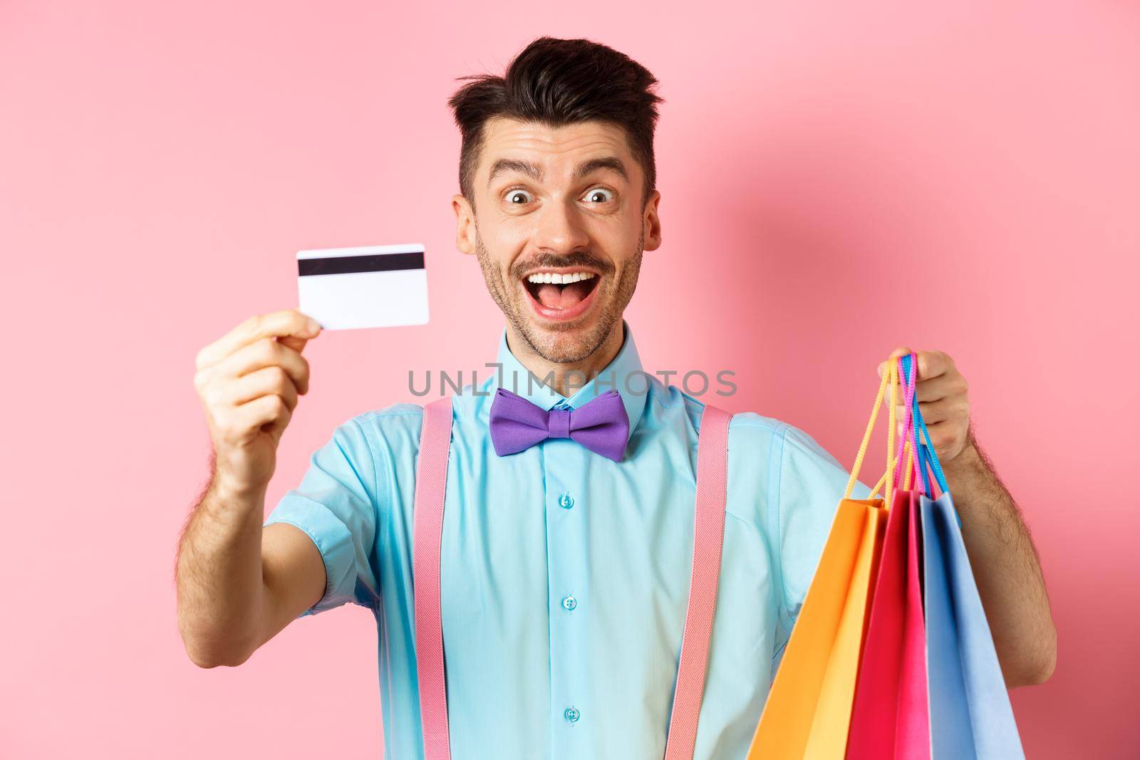 Happy young man giving his credit card and holding shopping bags, buying with discounts, standing on pink background and smiling by Benzoix