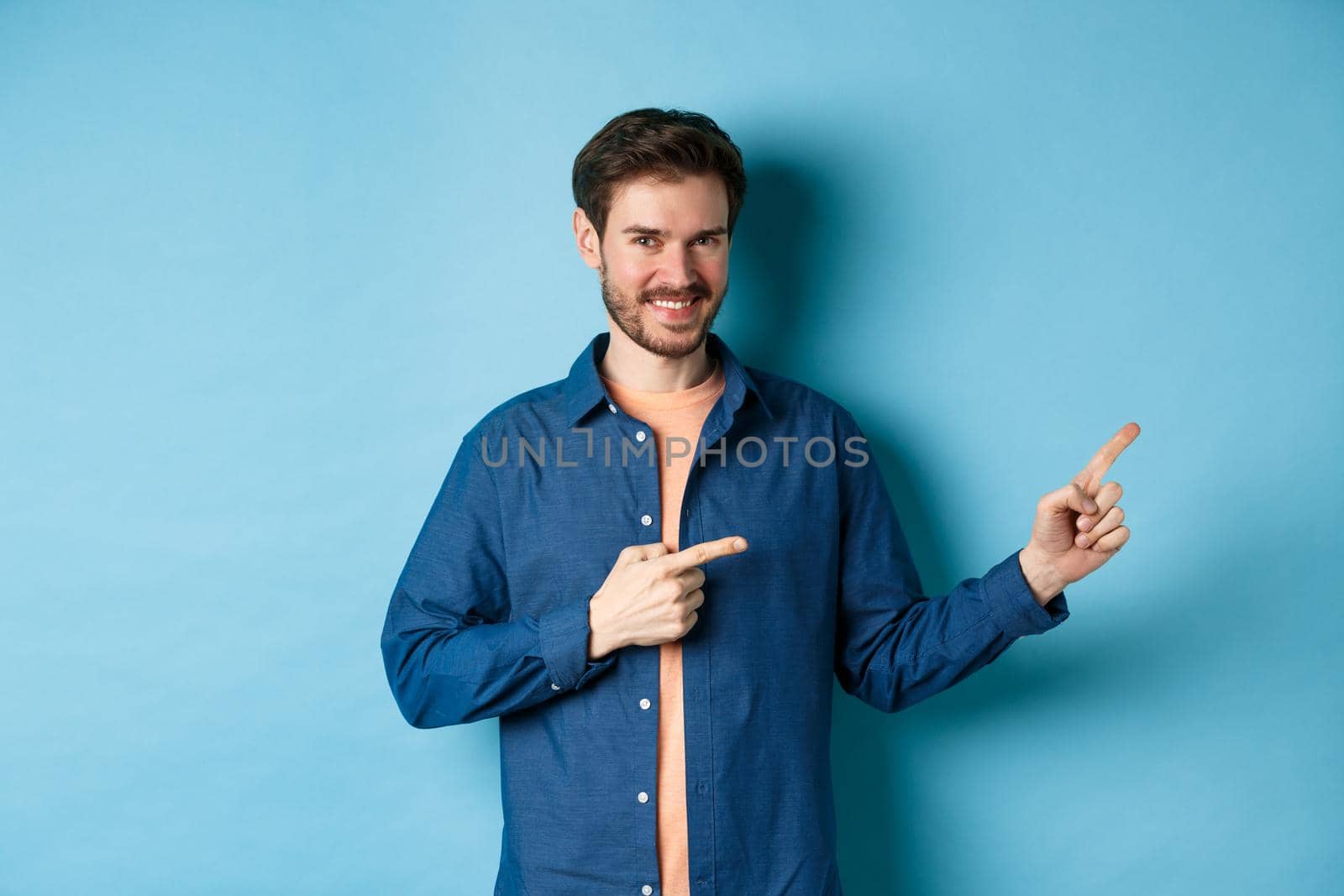 Smiling confident man showing logo, pointing fingers right at empty space and looking at camera, standing on blue background by Benzoix