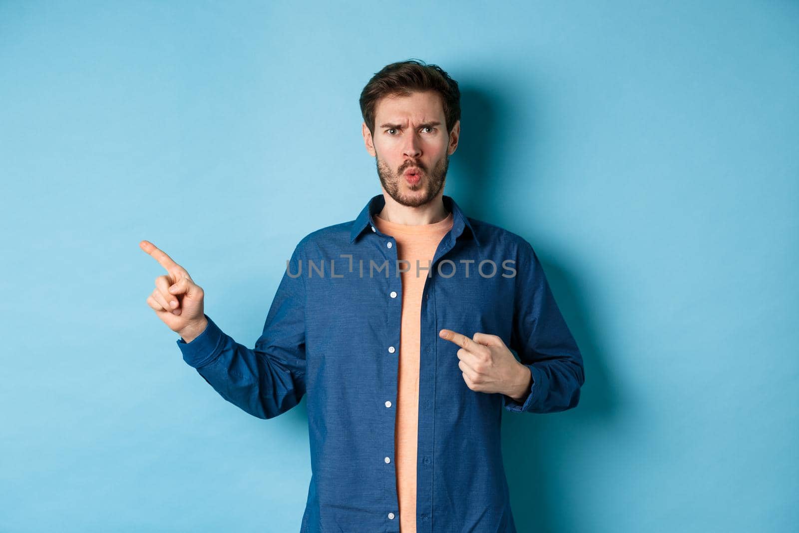 Shocked and offended young man frowning and looking hurt, pointing fingers left at empty space, complaining on something, standing on blue background by Benzoix