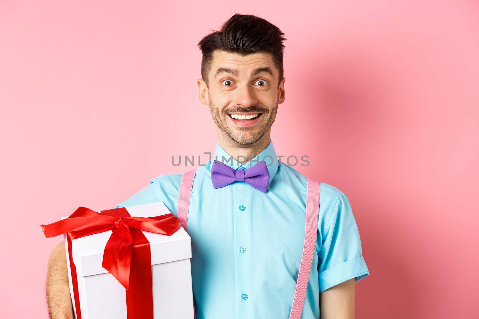 Holidays and celebration concept. Cheerful guy in bow-tie and suspenders bring gift box to party, holding present and smiling, standing over pink background.