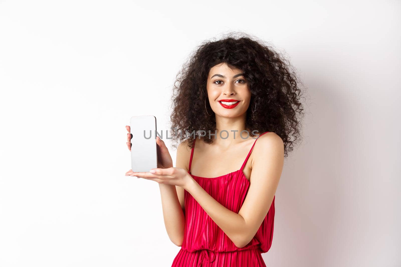 Elegant woman in red dress and makeup, showing blank smartphone screen and smiling, standing over white background. Copy space