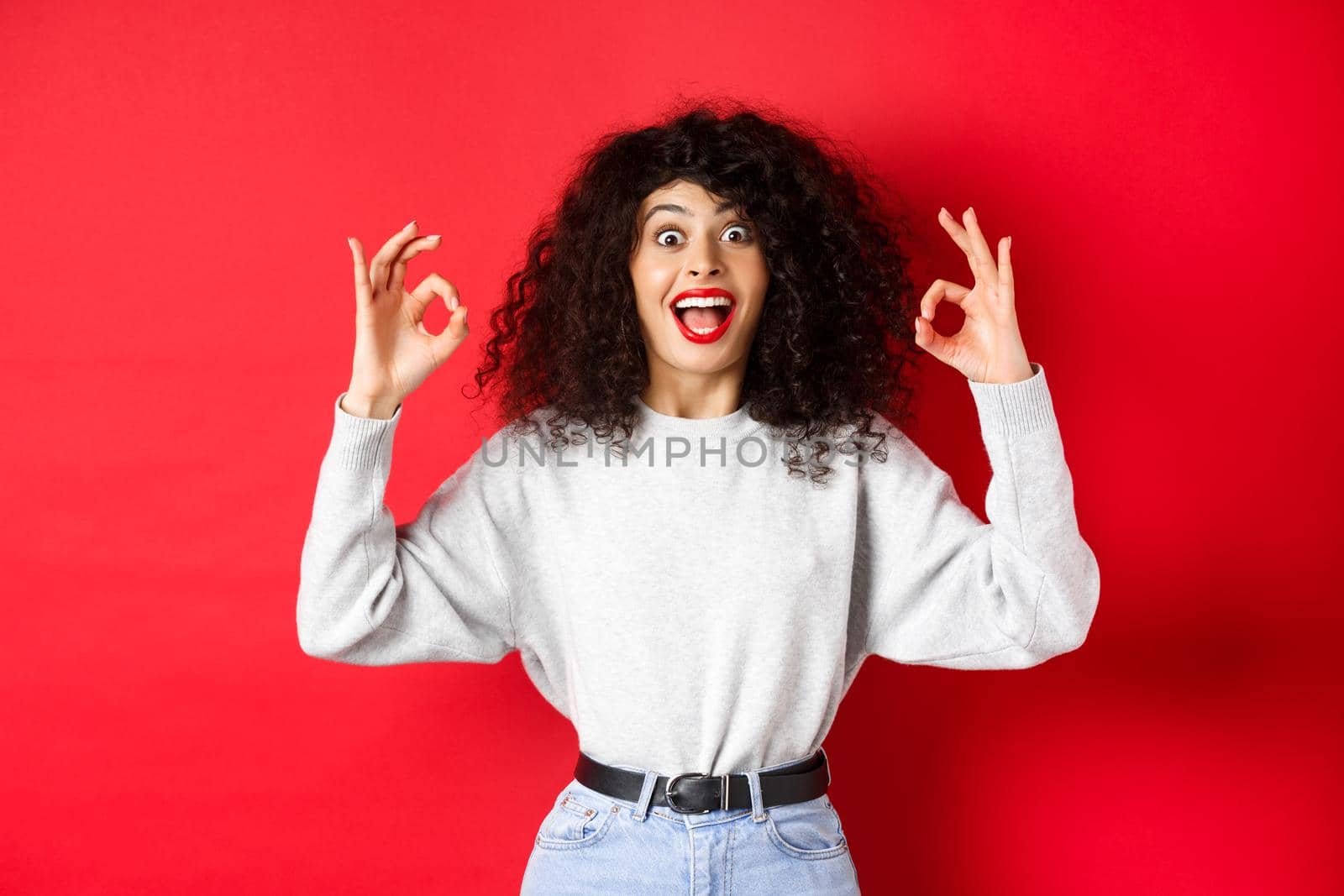Excited girl show okay signs and smiling happy, recommending good item, praising choice, standing amazed on red background.