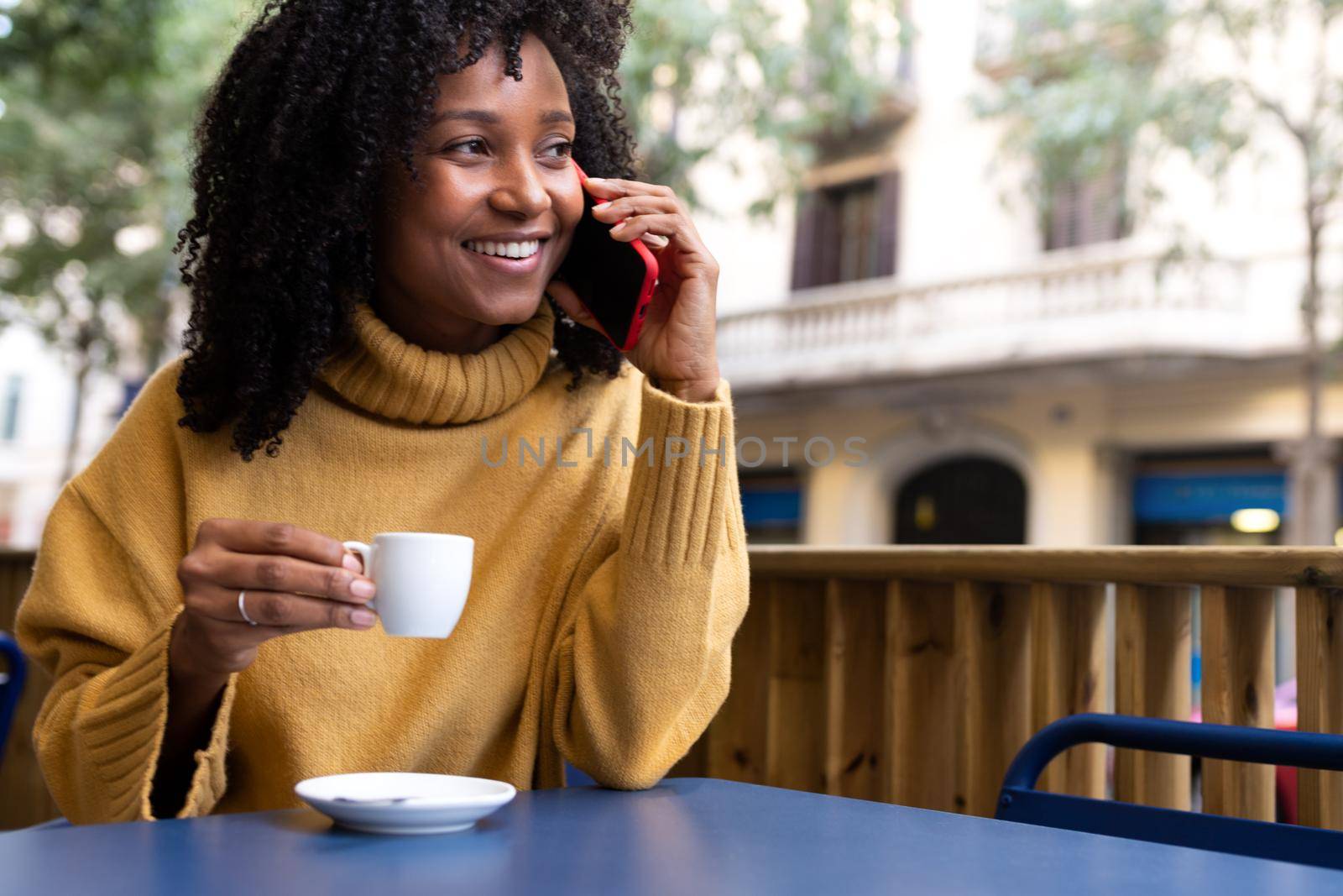 Smiling young African American woman talking on mobile phone and drinking espresso coffee at outdoors cafe terrace. Copy space. Lifestyle and technology concept.