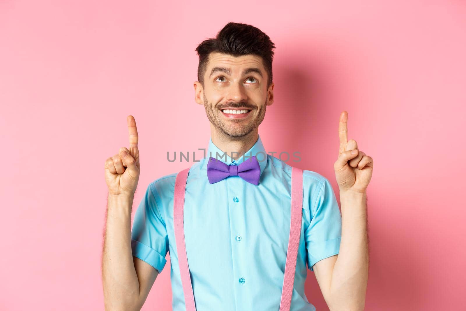 Happy smiling man in bow-tie pointing, looking up, showing something cool, standing on pink background.