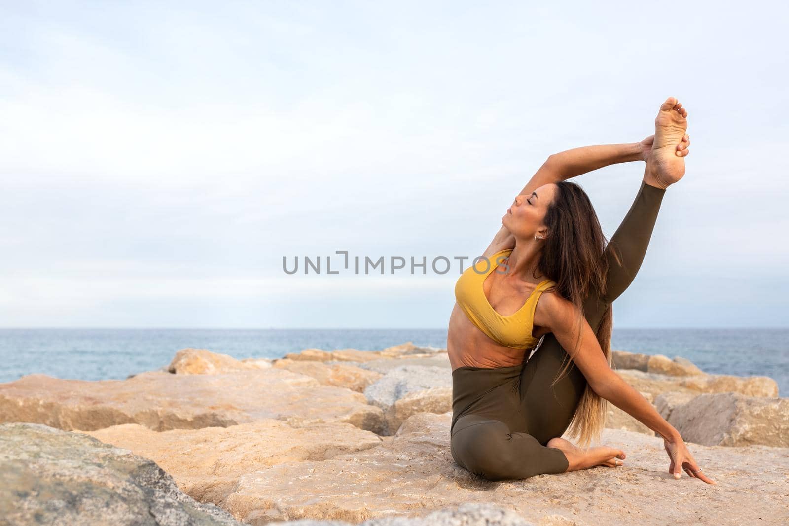 Flexible young woman doing Parivritta Kraunchasana yoga pose on a rock near the sea. Copy space. Healthy and active lifestyle concept.