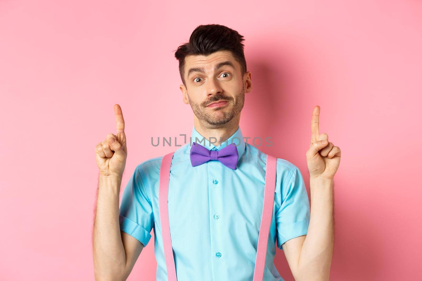 Cheerful funny man in bow-tie pointing fingers up, showing advertisement and smiling indecisive, standing over pink background. Copy space