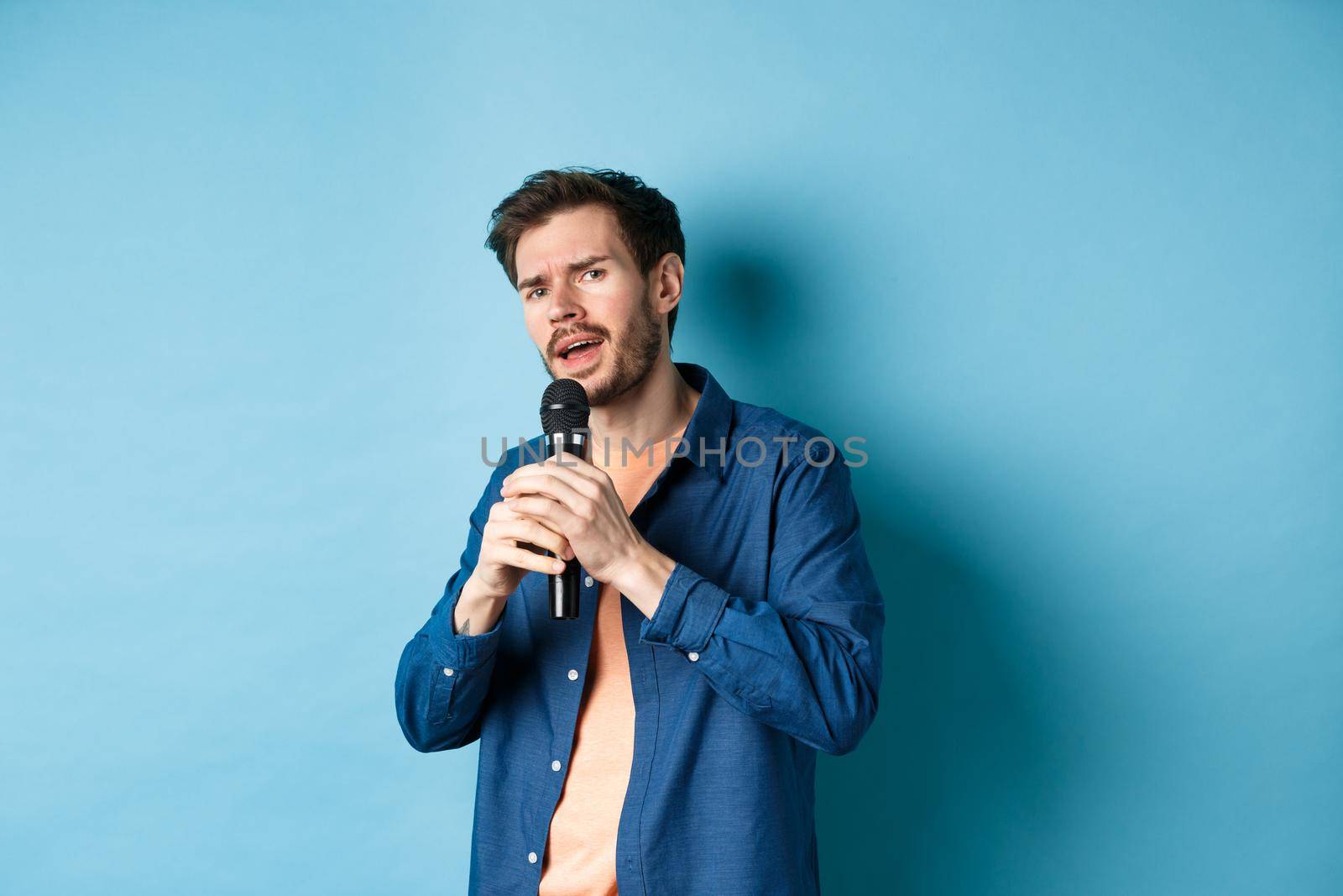 Young man singing song in microphone, playing karaoke, standing on blue background.