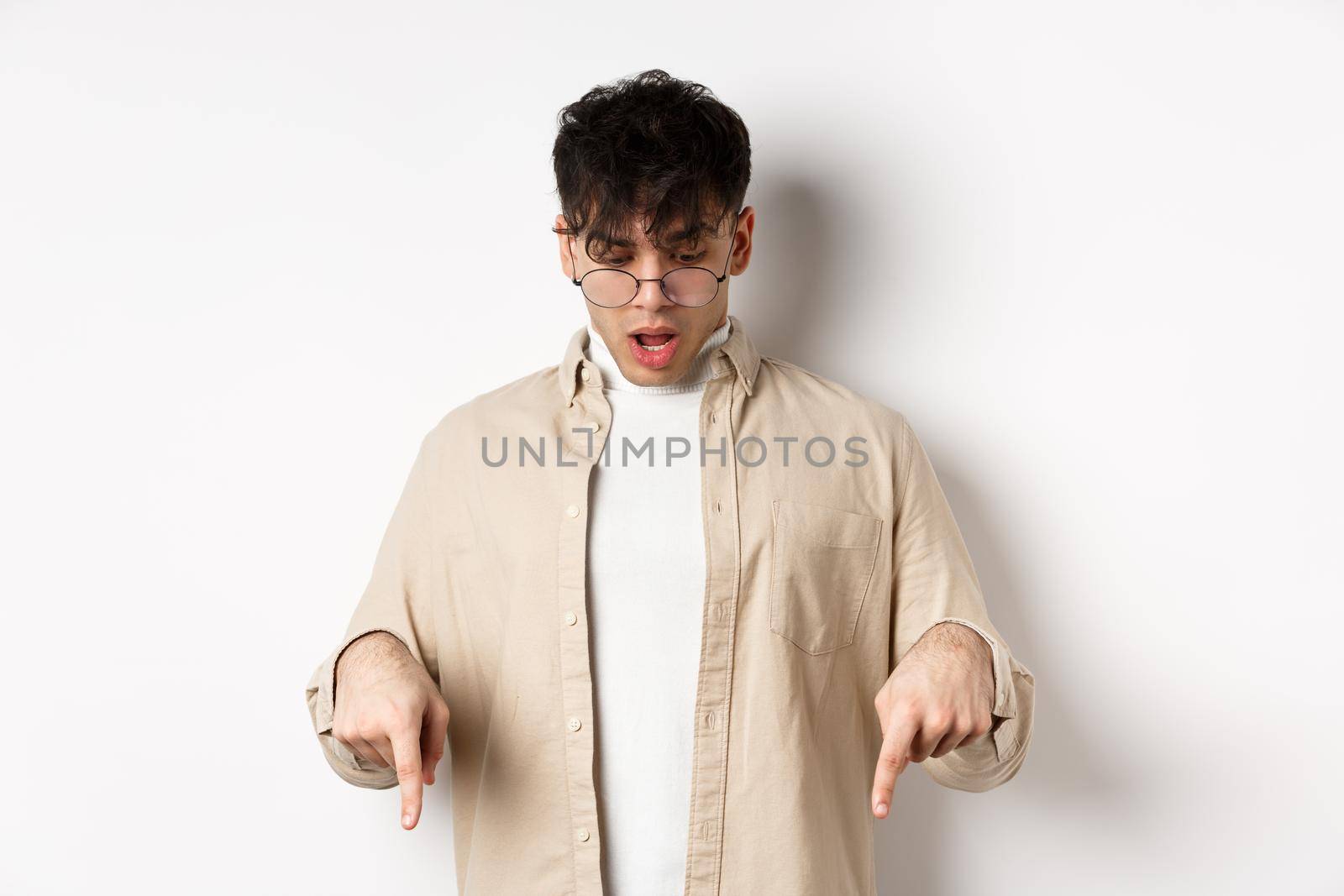 Amazed young guy in glasses look and point down, open mouth and say wow impressed, checking out discount offer, standing on white background.