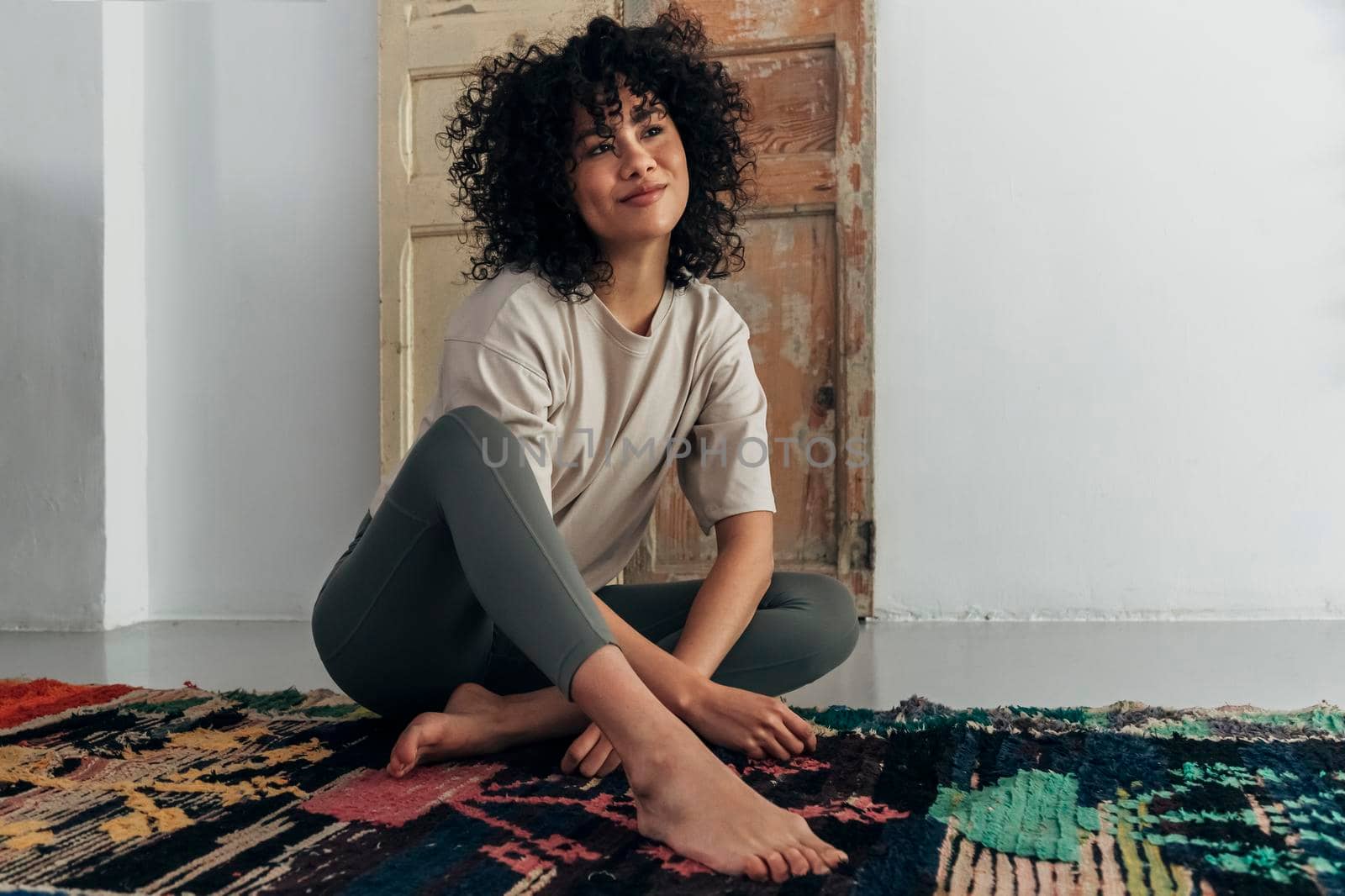 Pensive young multiracial smiling woman sits on floor looking away. Copy space. by Hoverstock