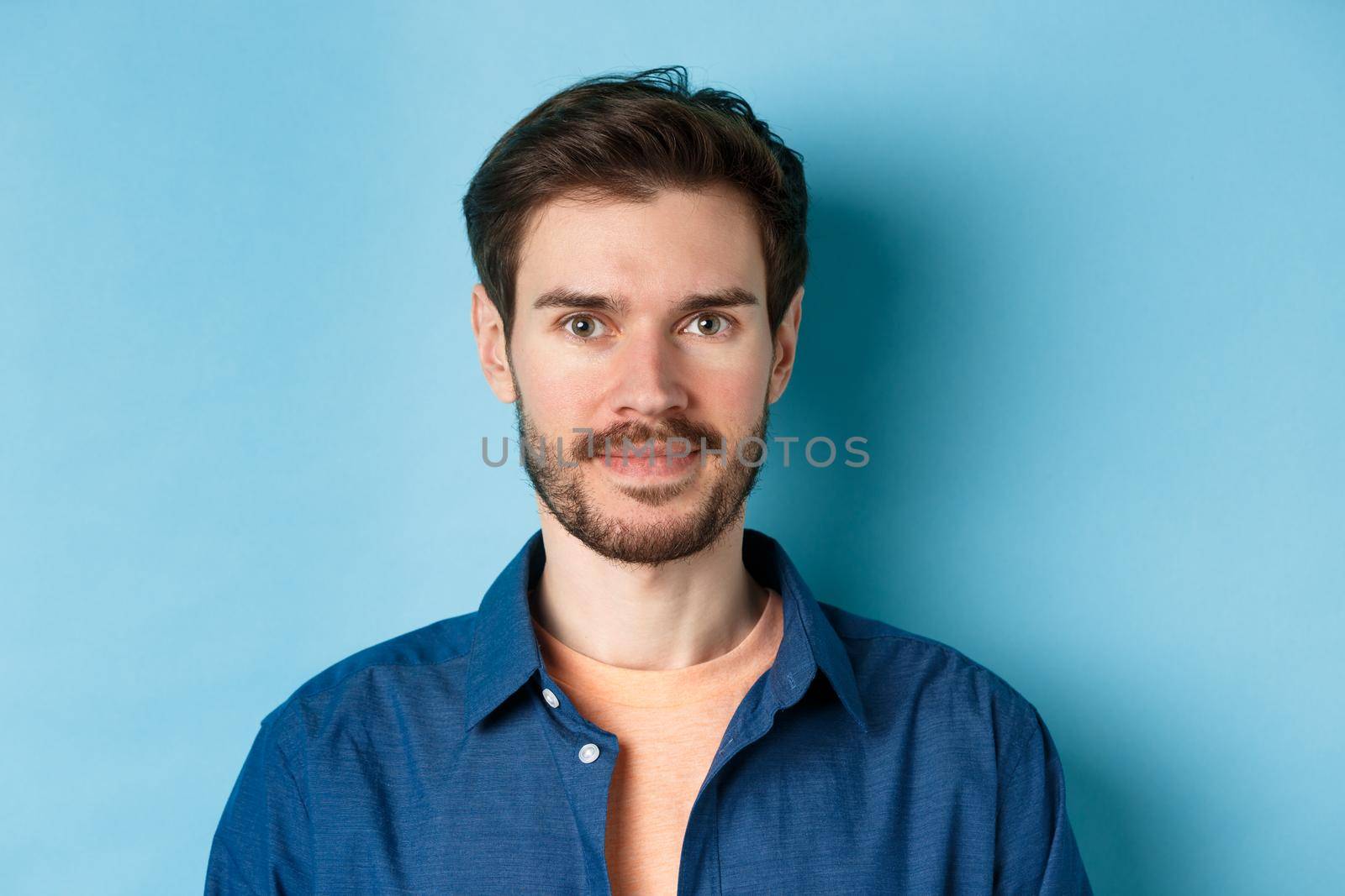 Close-up of young caucasian guy with beard smiling at looking happy at camera, standing on blue background by Benzoix