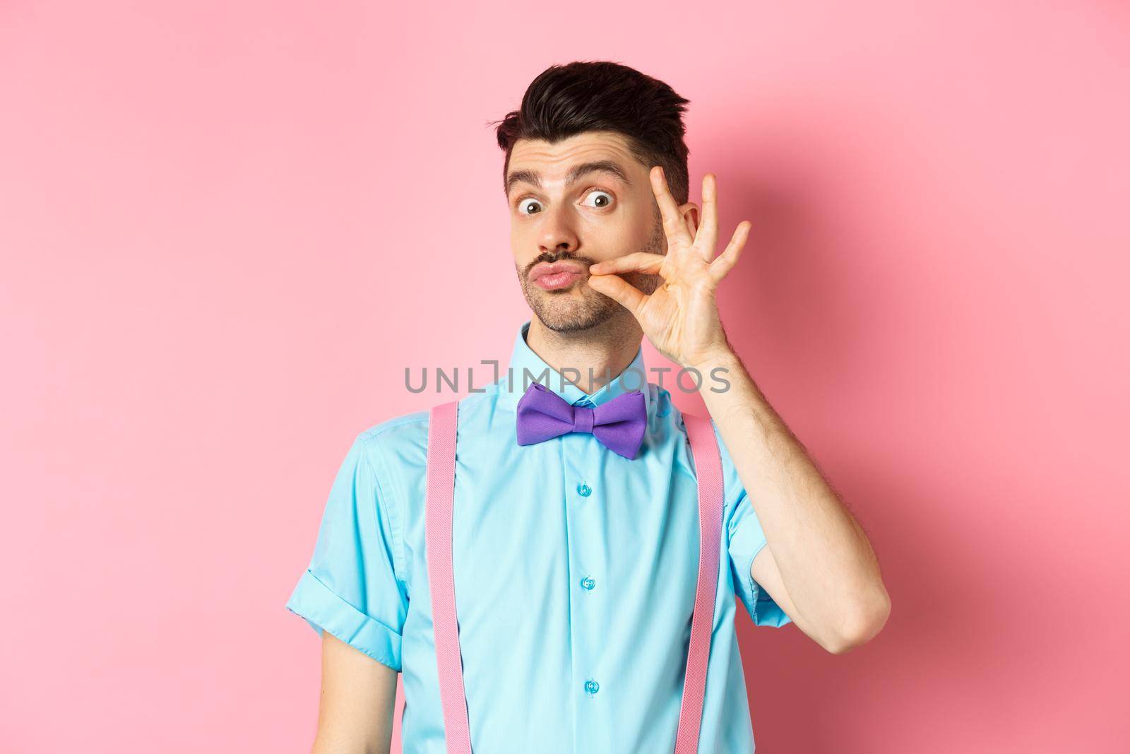 Funny young man touching his french moustache and pucker lips, looking silly at camera, standing in bow-tie and suspenders on pink background by Benzoix