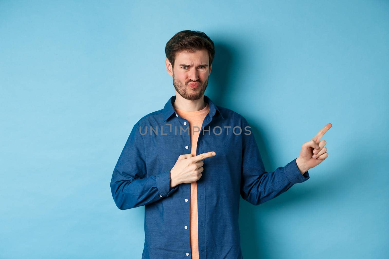 Disappointed young man frowning and pointing fingers right at empty space, looking upset and displeased, complaining, standing on blue background by Benzoix