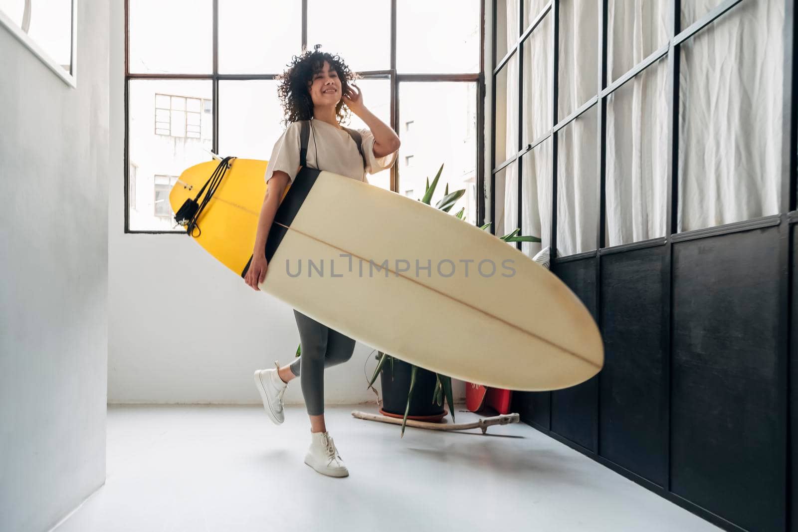 Smiling young mixed race woman arrives home listening to music and carrying a surf board after a day of surfing. Lifestyle concept.