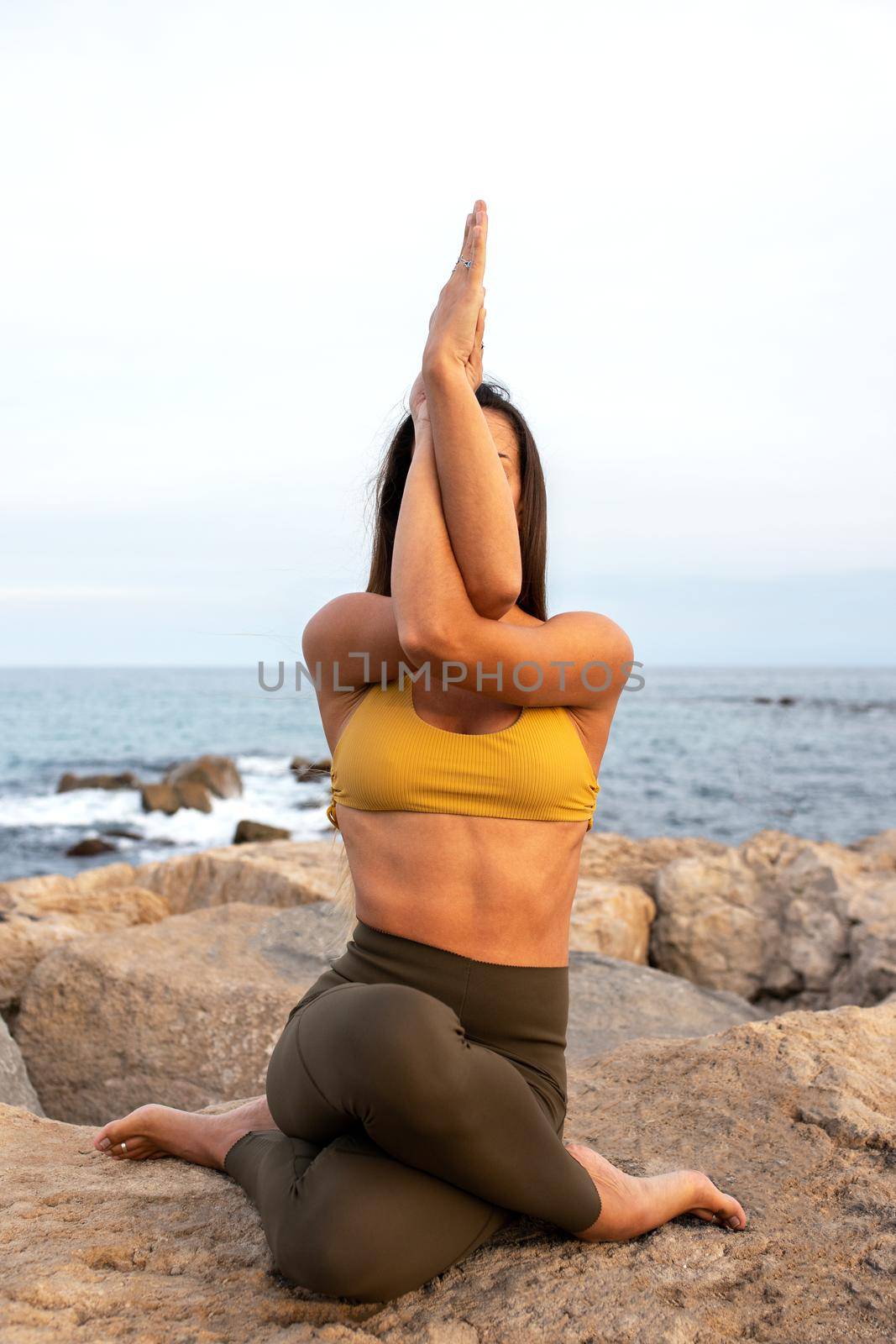 Unrecognizable woman doing gomukhasana with garudasana arms. Yoga practice in nature near the ocean. Vertical image. Healthy lifestyle and spirituality concept.