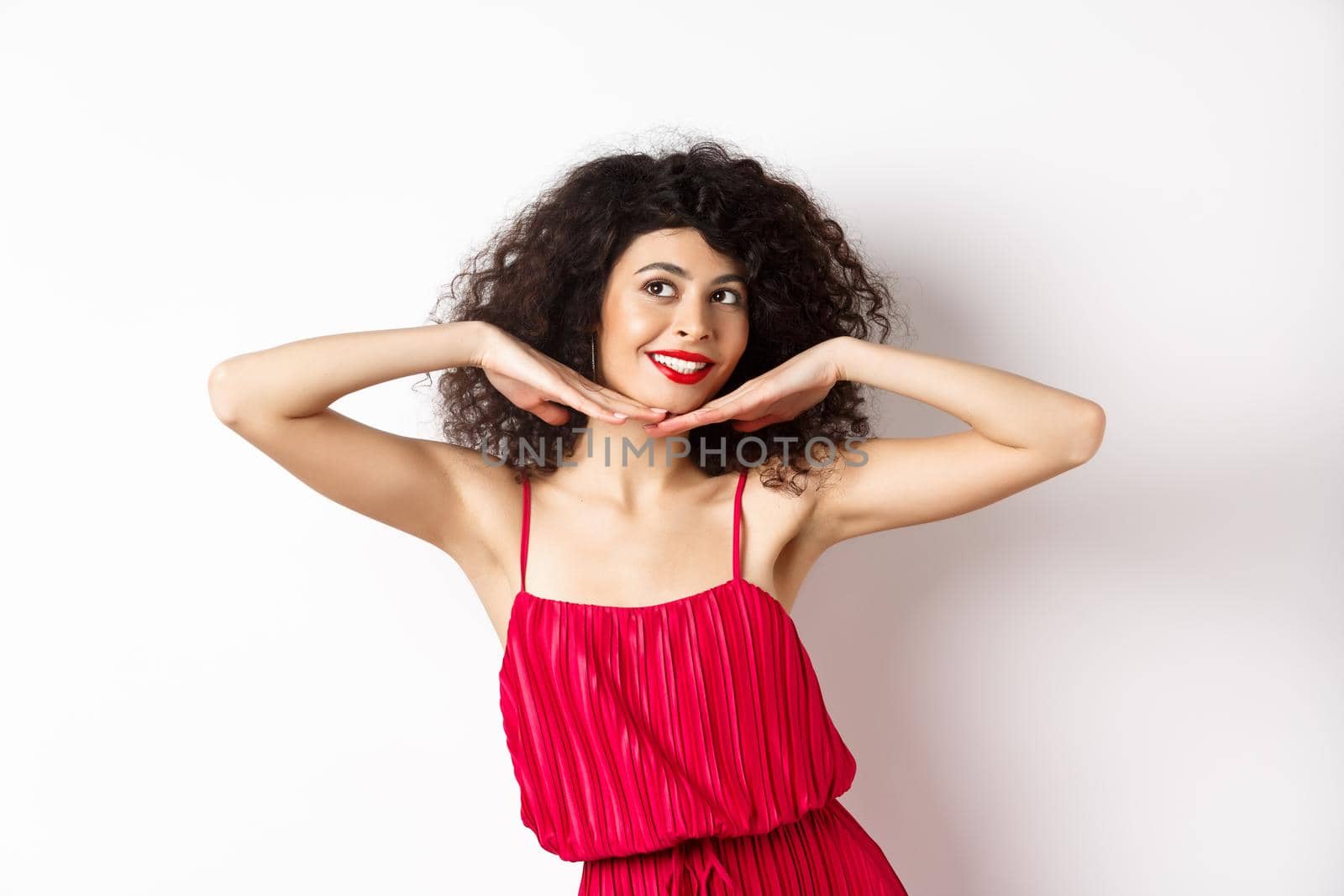 Beautiful lady with curly hair, wearing red dress, showing hear face with makeup and smiling, feeling carefree on white background.