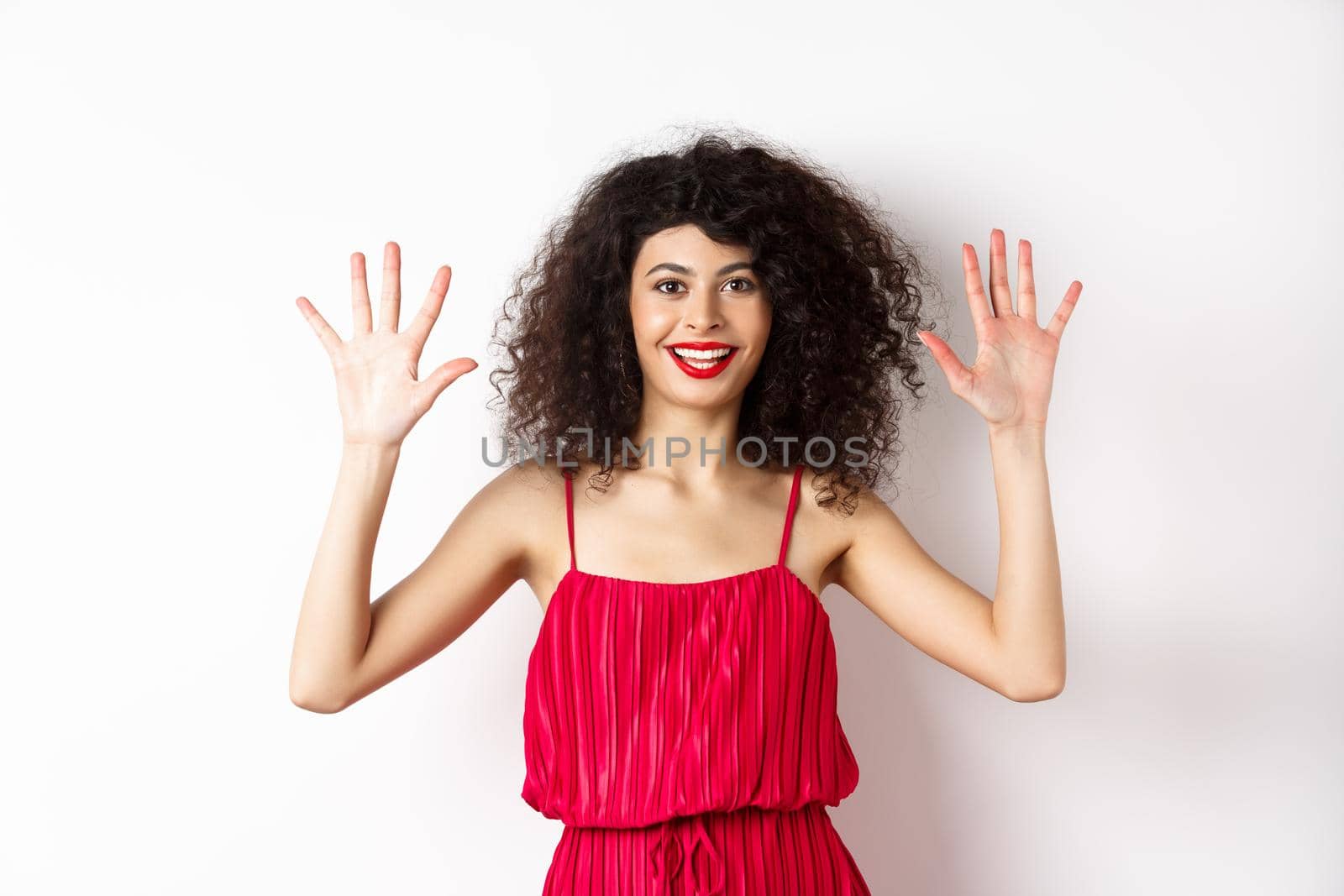 Beautiful female model with curly hair, wearing red dress, showing number ten with fingers, standing against white background.