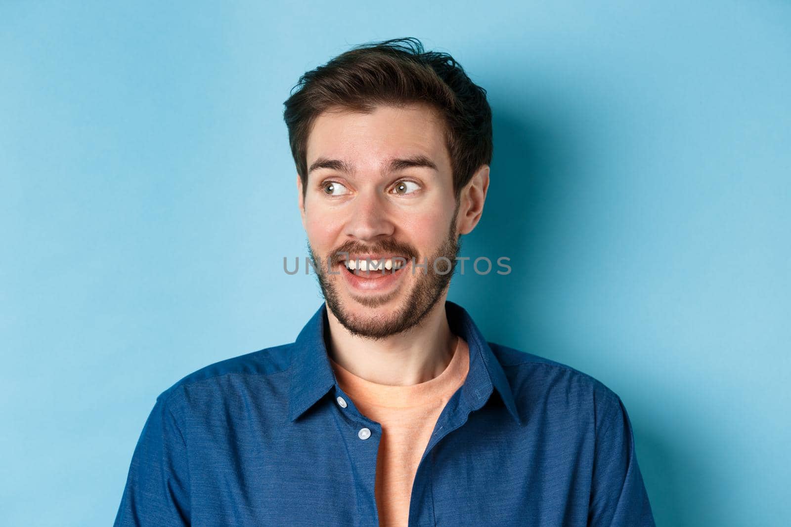 Close-up of cheerful bearded guy looking left at empty space and smiling, standing happy on blue background.