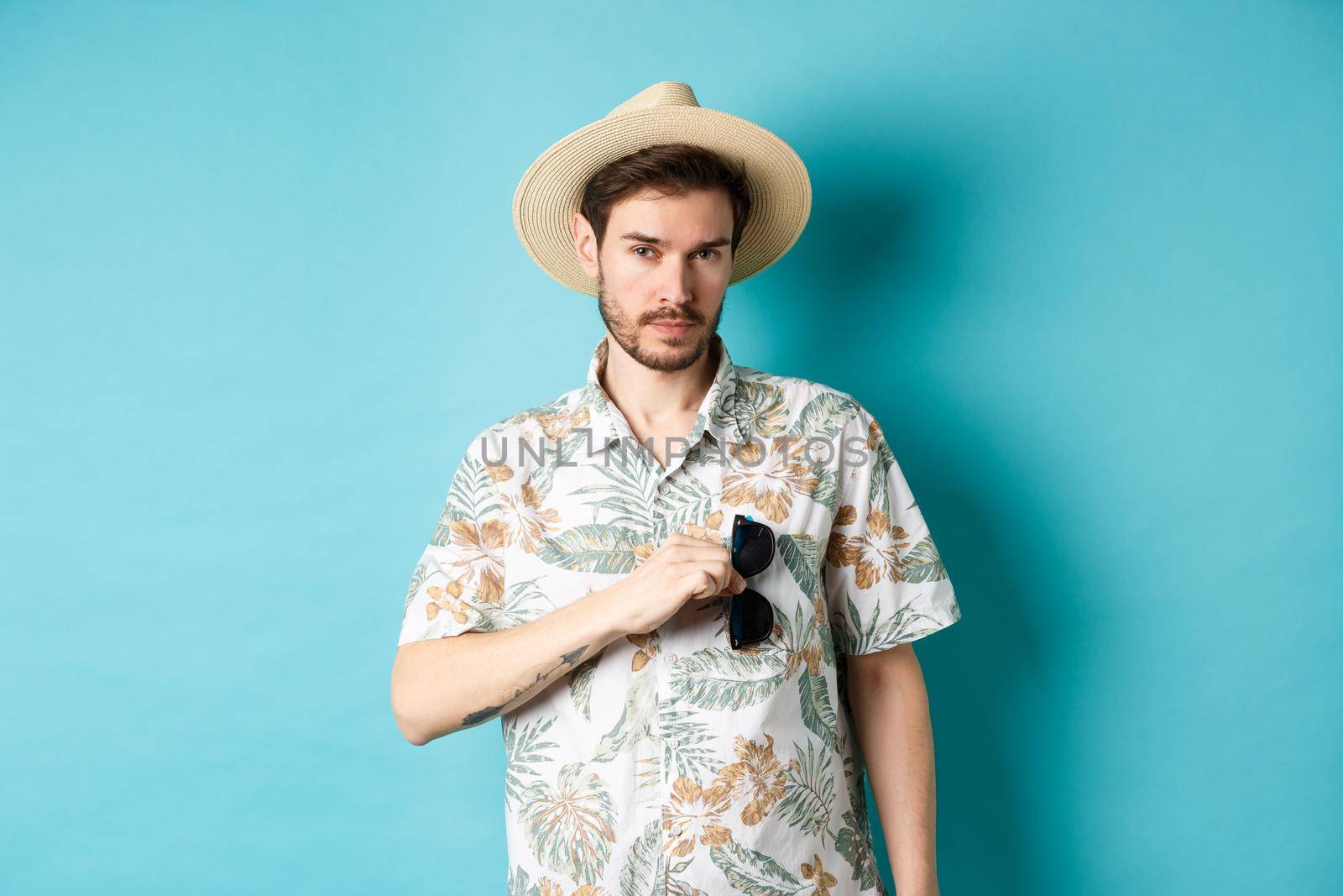 Handsome tourist in hawaiian shirt and summer hat, put sunglasses in pocket, going on vacation, standing against blue background.