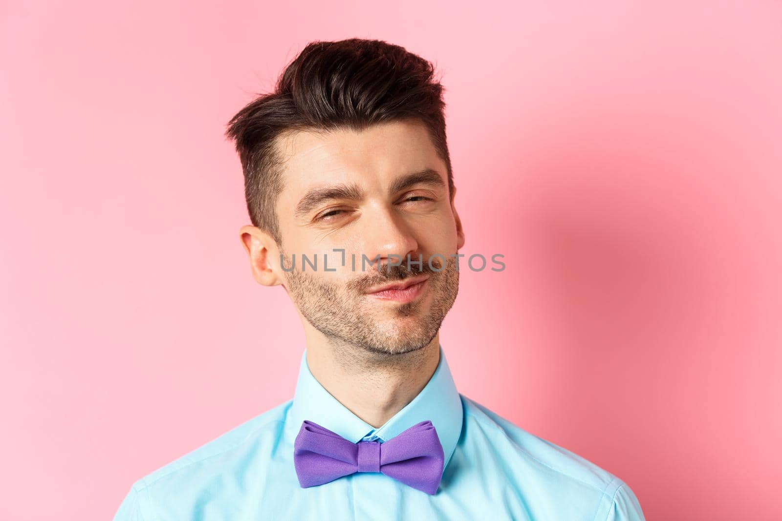 Close-up of smiling young guy with moustache, squinting intrigued, listening to something interesting, standing in bow-tie on pink background.