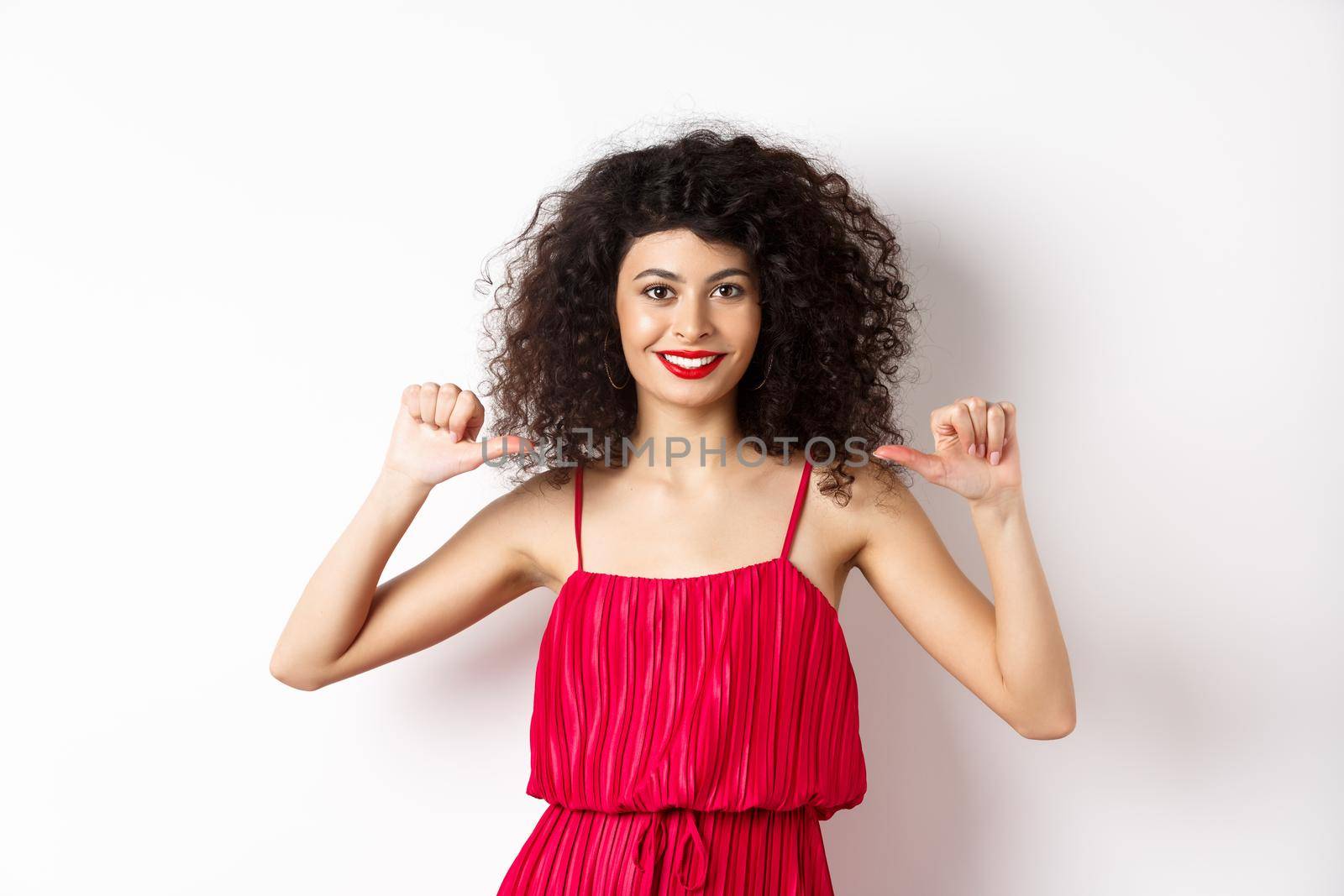 Confident young woman in elegant red dress, pointing at herself and smiling, self-promoting, standing over white background.
