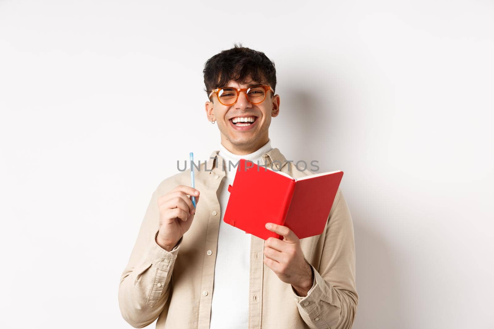 Cheerful young man in glasses laughing and taking notes, writing down in planner, holding pen and diary, standing on white background by Benzoix