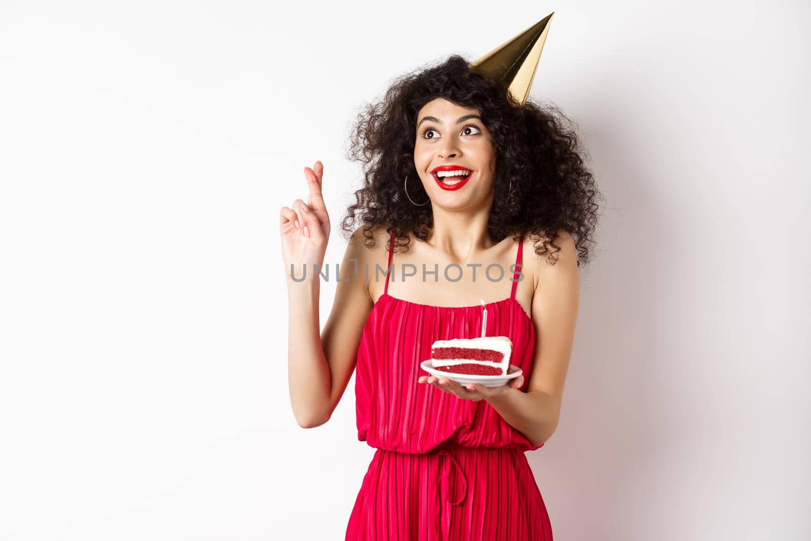 Excited birthday girl making wish, cross fingers good luck and looking aside at logo, celebrating bday, holding piece of cake, white background by Benzoix