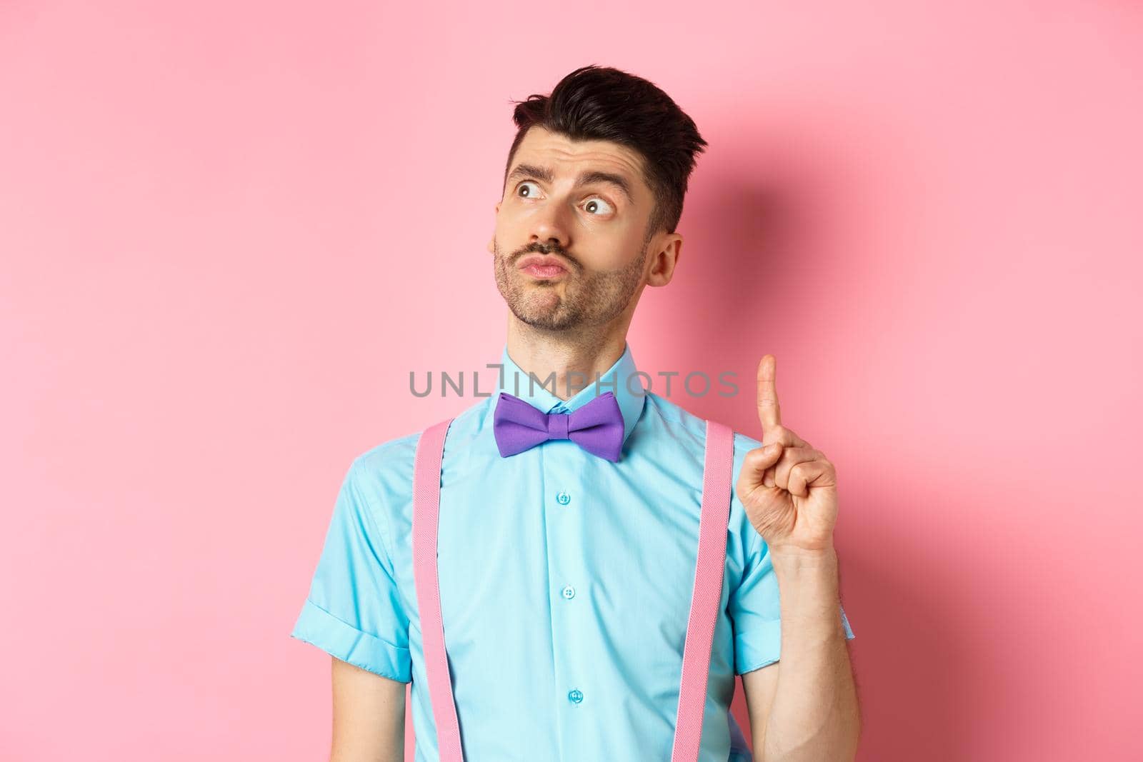 Pensive handsome man in bow-tie pitching an idea, raising index finger while looking away with thoughtful face, standing on pink background.