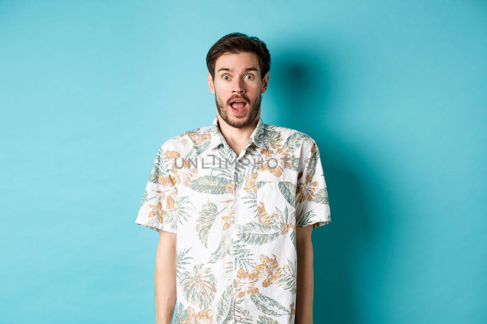Summer holiday. Surprised guy in hawaiian shirt gasping amazed, checking out vacation offer, standing on blue background.