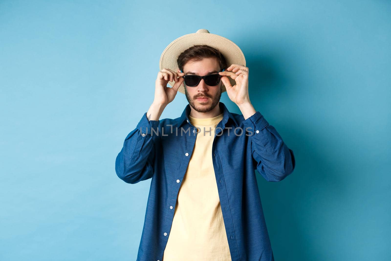 Handsome tourist in straw hat put on sunglasses on summer vacation, standing on blue background.