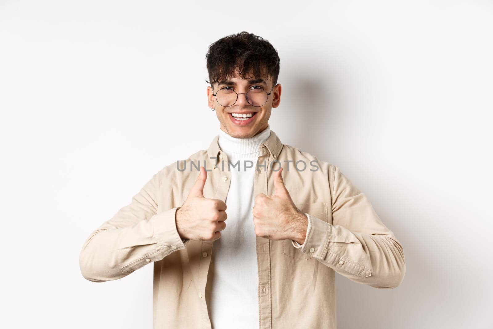 Cheerful hipster guy in glasses showing thumbs up and smiling, like good product, recommend or praise advertisement, standing on white background.