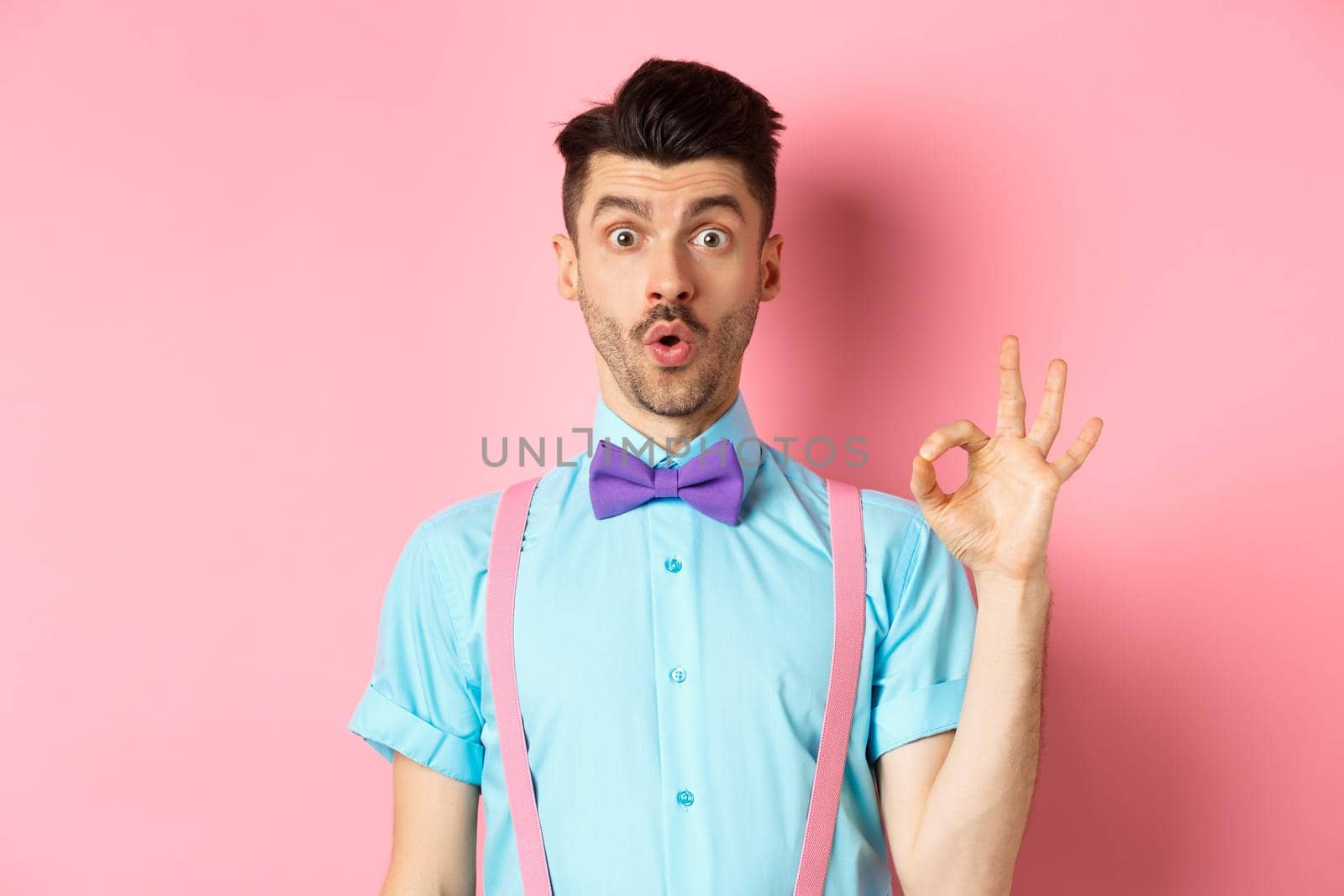 Impressed guy in bow-tie and suspenders saying wow, showing okay gesture with amazement, praise awesome thing, standing over pink background by Benzoix