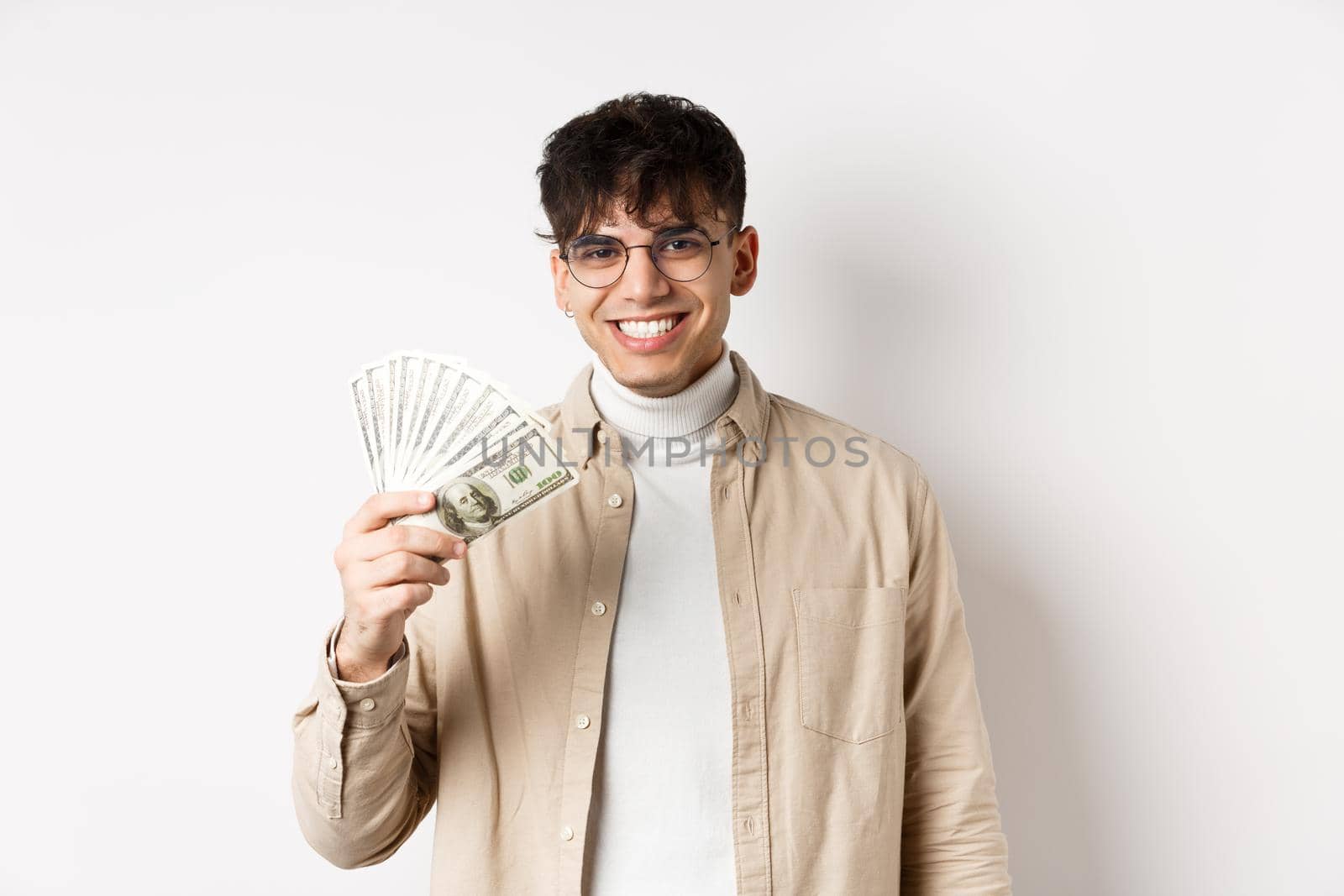 Happy young guy holding dollar bills and smiling, making money and looking cheerful at camera, standing on white background.