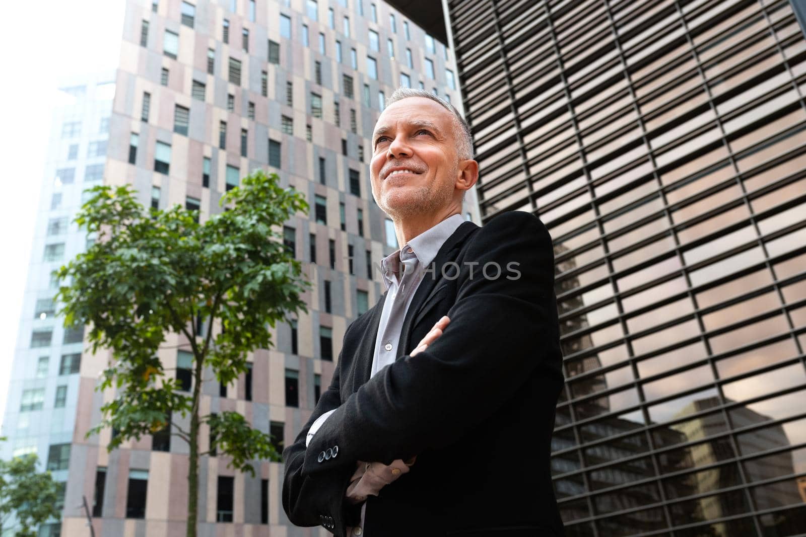 Pensive business man stands next to buildings with arms crossed. Copy space. by Hoverstock
