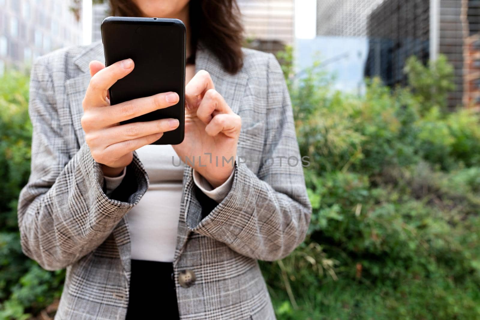 Unrecognizable young business woman using mobile phone near office buildings. Selective focus on hands and phone. Copy space. Business and technology concept.