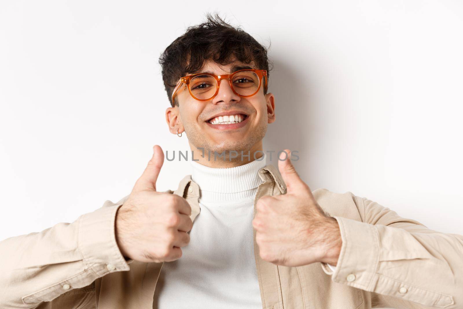 Close-up of satisfied happy guy showing thumbs up and smiling with white teeth, wearing eyewear, standing on white background.