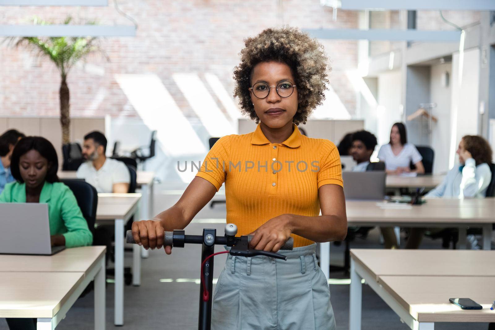 Portrait of young african american woman looking at camera with blonde curly hair leaving the office with electric scooter. Sustainable lifestyle concept.