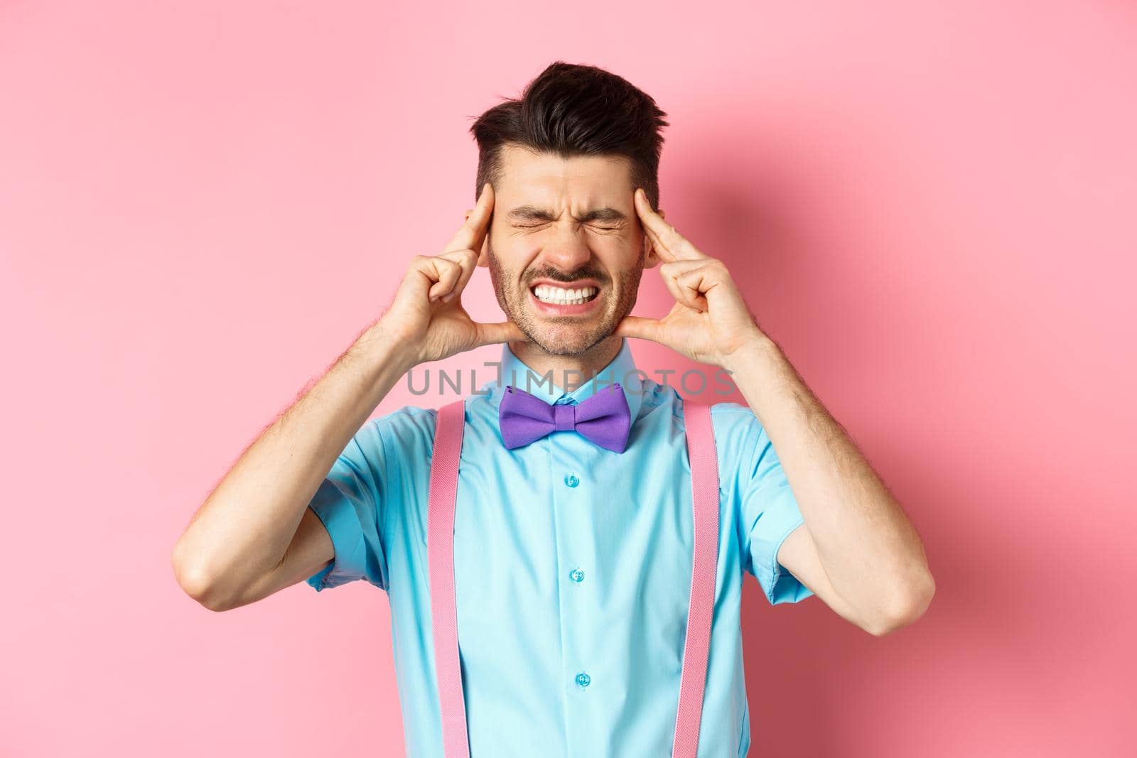 Image of man with headache touching head temples, grimacing from painful migraine, clench teeth, standing sick on pink background.