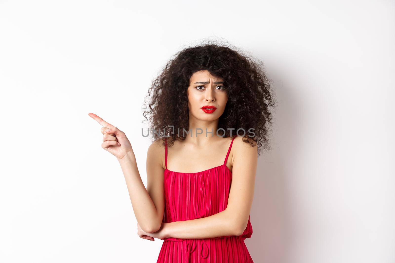 Arrogant young woman with curly hair, wearing red dress, frowning and complaining, pointing finger left at promo, standing over white background.