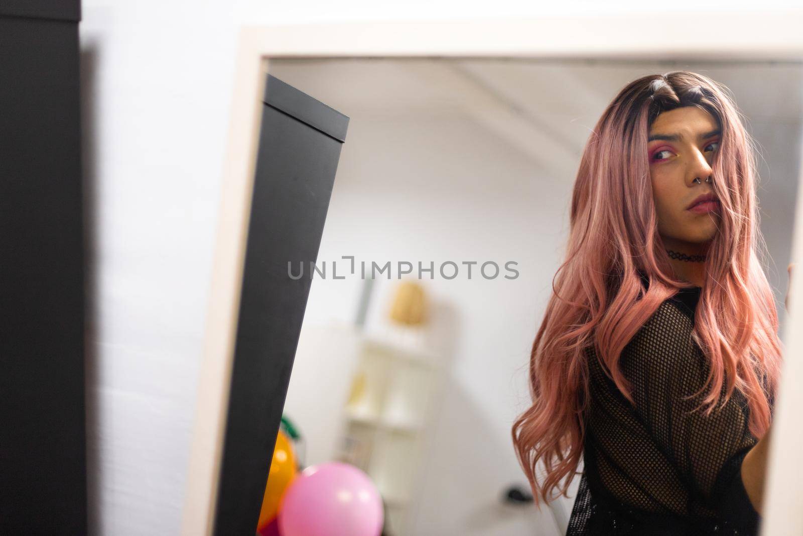 Young caucasian man wearing long pink wig and makeup looking at himself in the mirror. Copy space. by Hoverstock