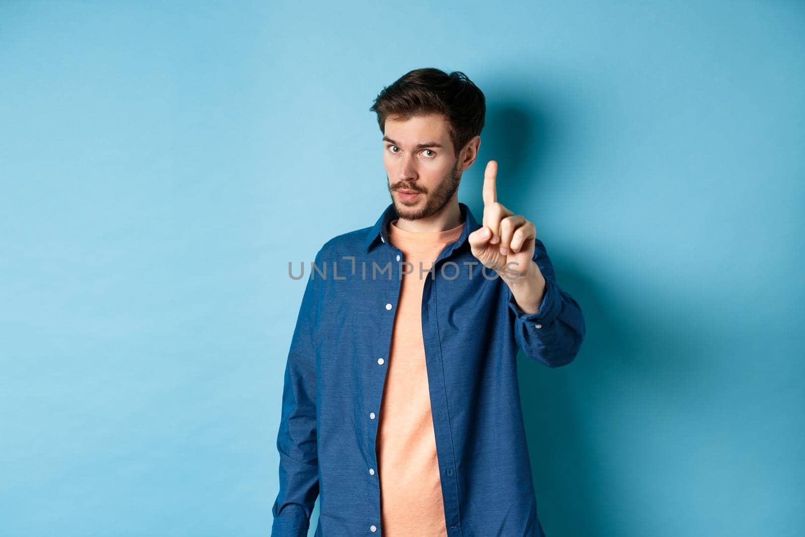 Image of young man give warning, teaching a lesson, raising one finger to scold, looking at camera, patronizing someone, standing on blue background.