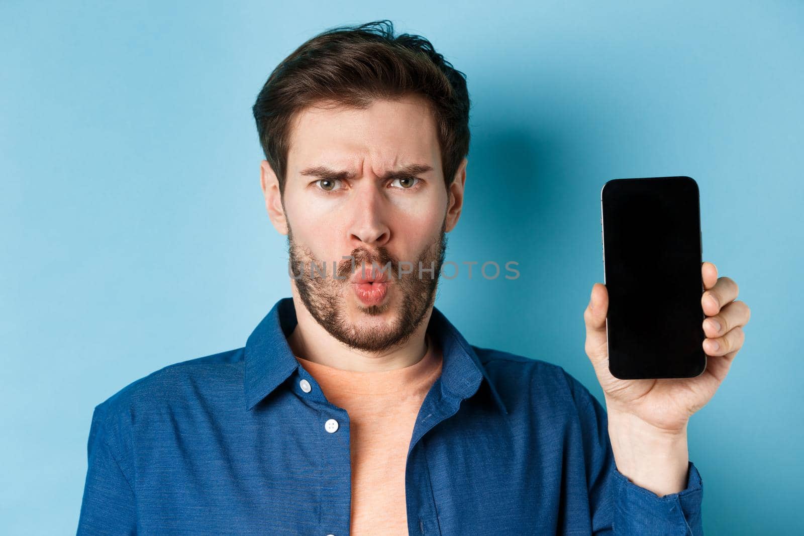 Shocked and confused young man frowning bothered, complaining at something on mobile phone, showing empty screen, standing on blue background.