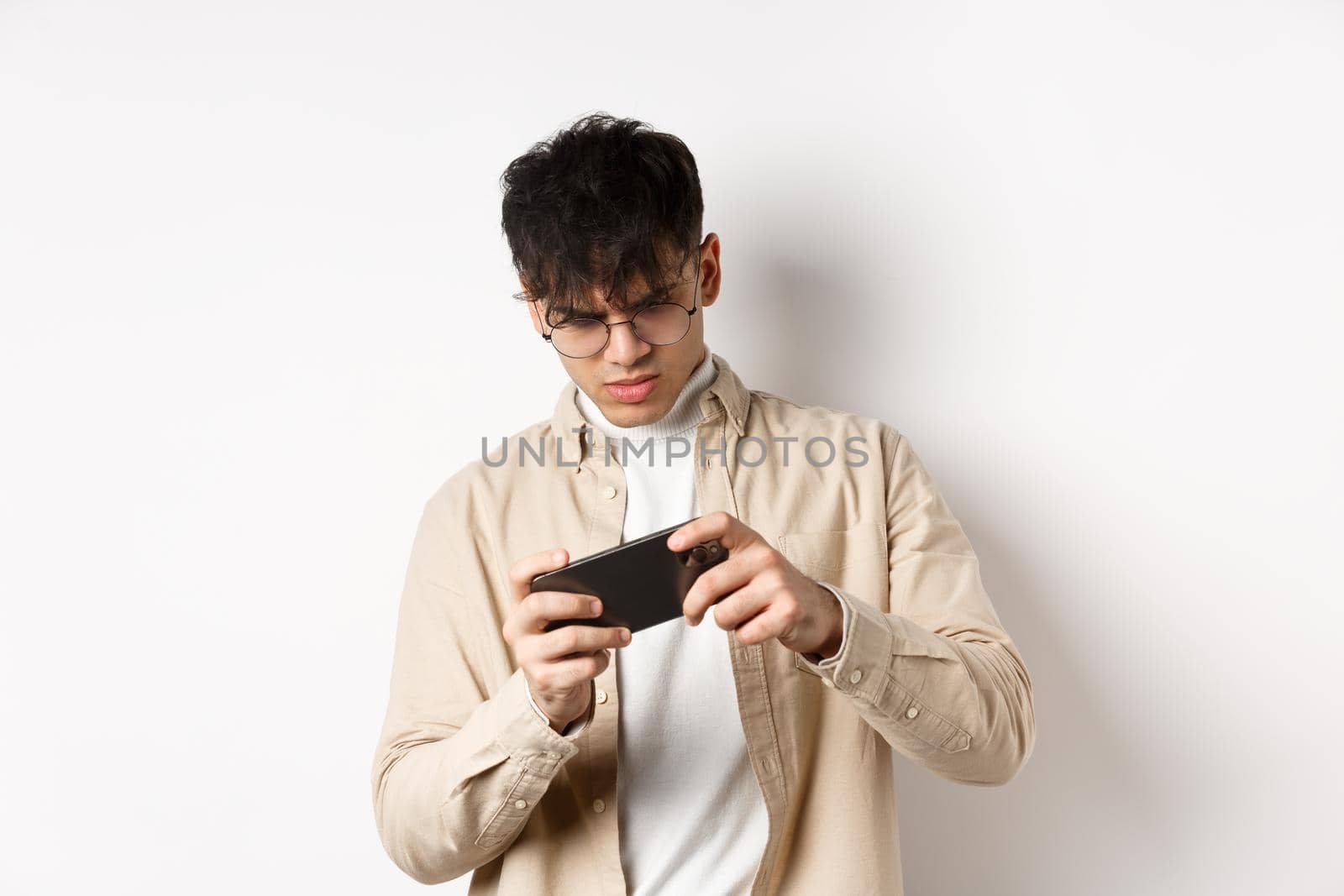 Portrait of handsome young man playing video game on mobile phone, tilt smartphone to play racing, standing on white background.